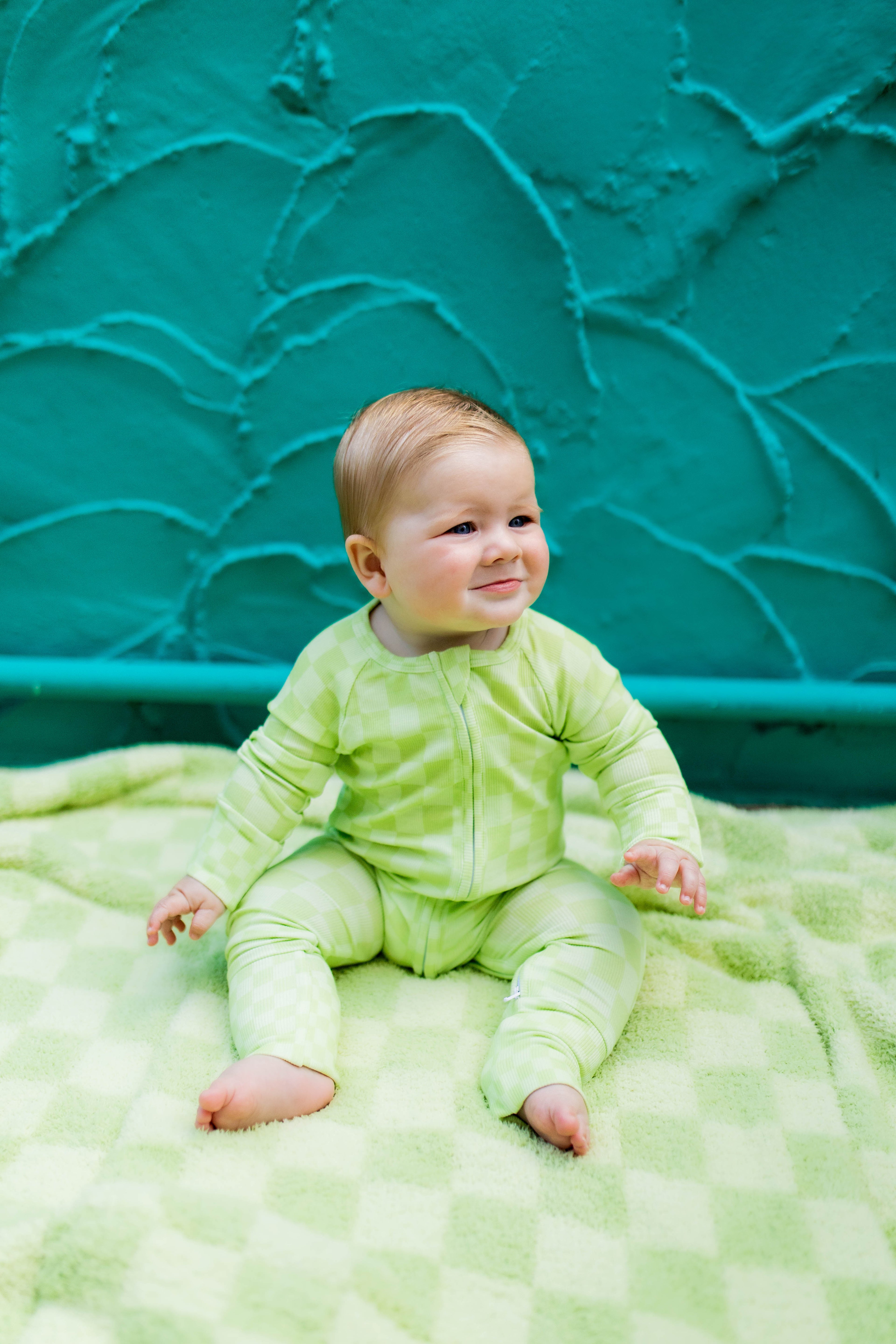 A baby wearing a light green long-sleeve romper sits on a green checkered blanket in front of a textured teal wall.