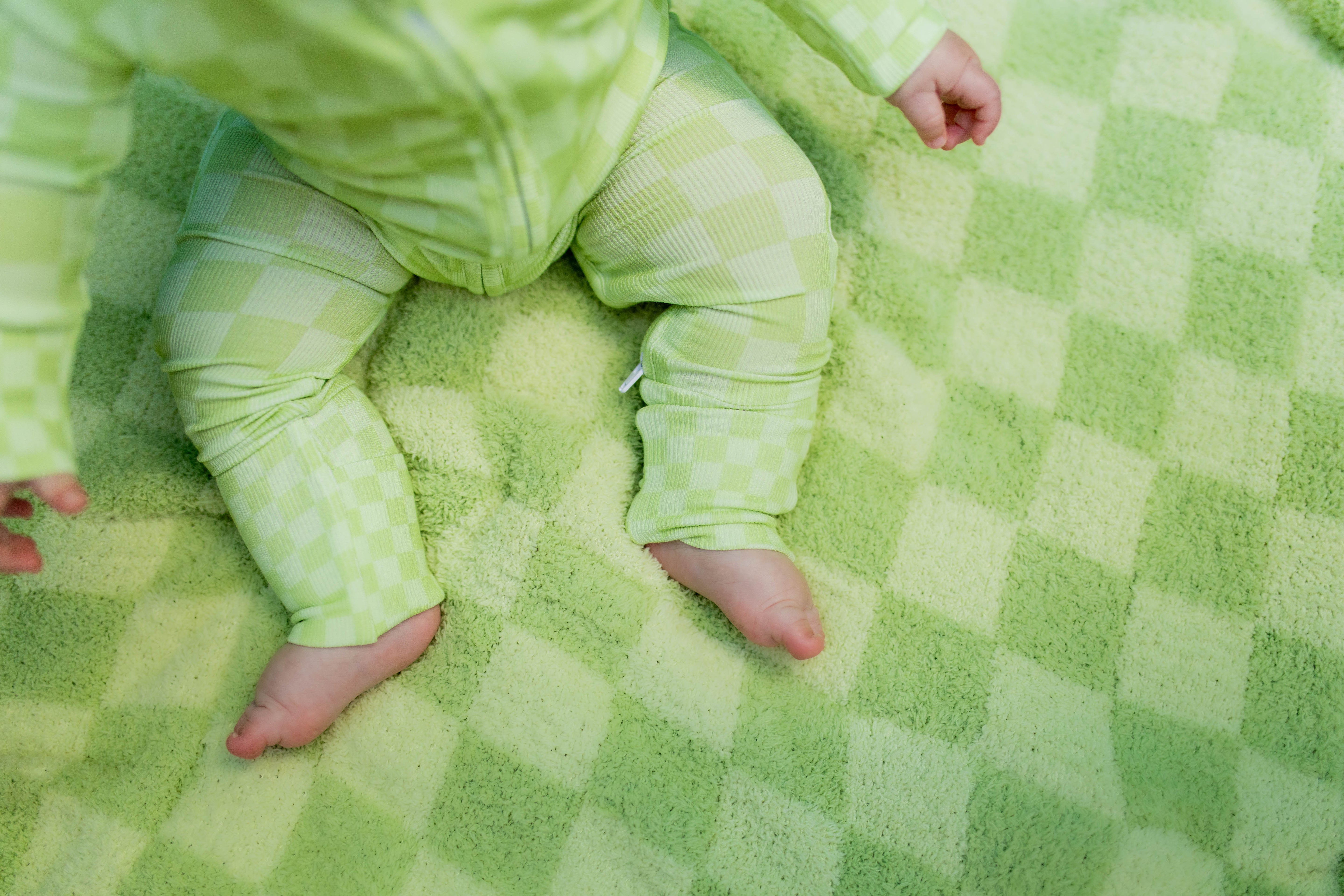 A baby wearing a light greencheckered one-piece outfit sits on a matching blanket.