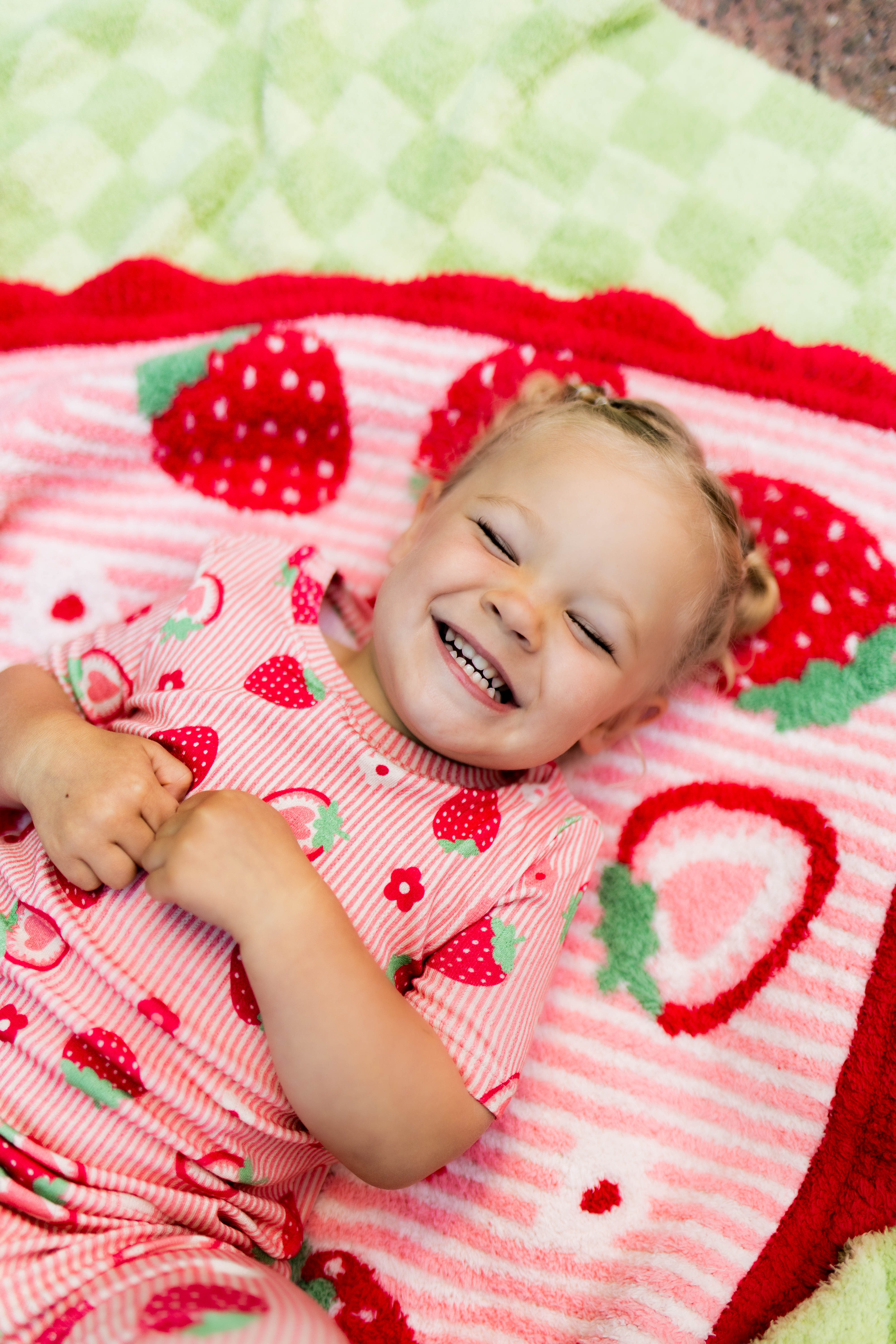 A smiling toddler lying on a strawberry-themed blanket, wearing a matching pink strawberry-print pajama top. The scene is styled with a cozy, playful aesthetic.