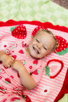 A smiling toddler lying on a strawberry-themed blanket, wearing a matching pink strawberry-print pajama top. The scene is styled with a cozy, playful aesthetic.