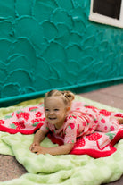 A smiling young girl lies on a light green plush blanket with a red and white strawberry-patterned blanket on top of it posed in front of a textured teal wall.