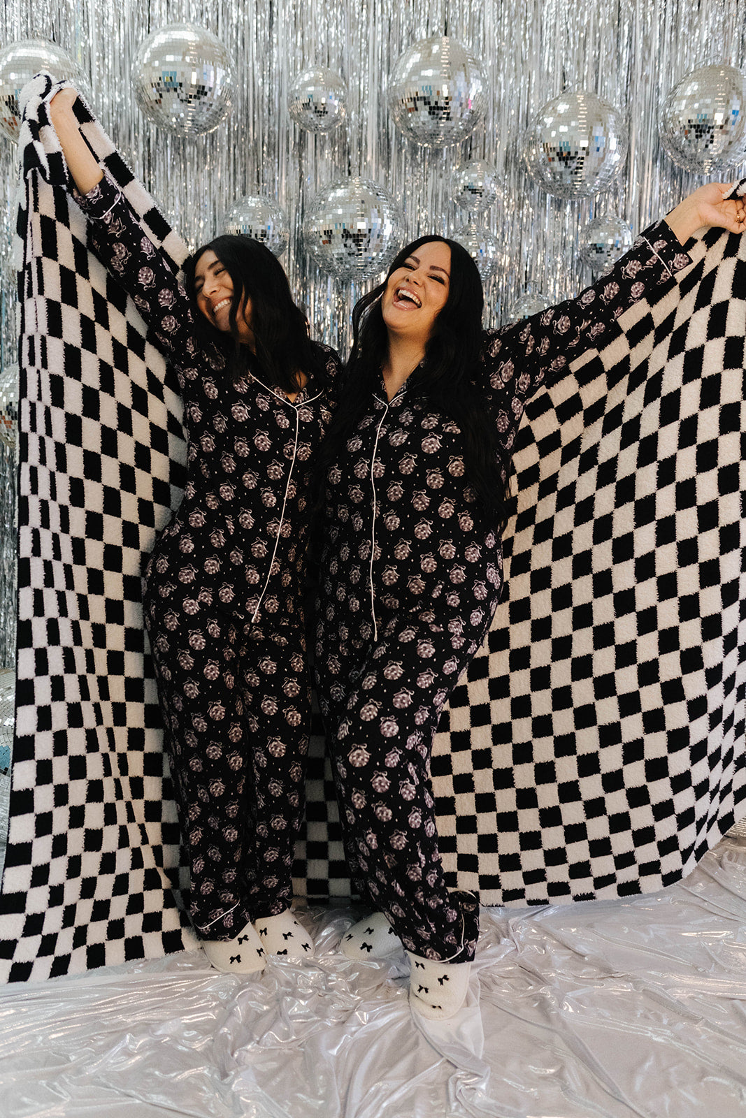 Two women holding a black and white checkered blanket against a silver disco ball background