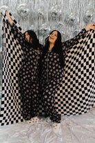 Two women holding a black and white checkered blanket against a silver disco ball background