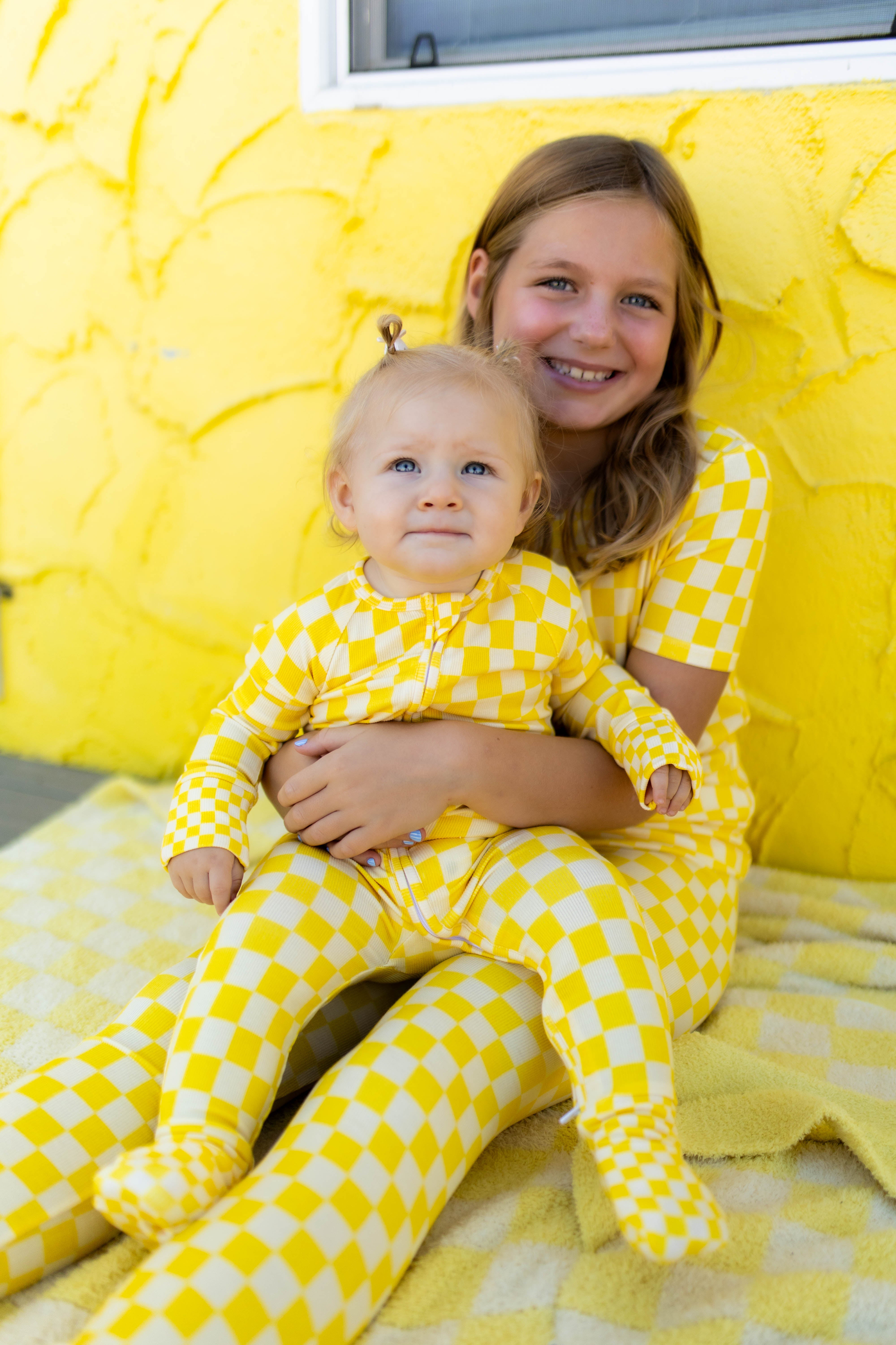 A smiling girl and a baby both wearing matching yellow checkered pajamas, posed together against a bright yellow textured wall.