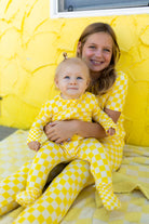 A smiling girl and a baby both wearing matching yellow checkered pajamas, posed together against a bright yellow textured wall.