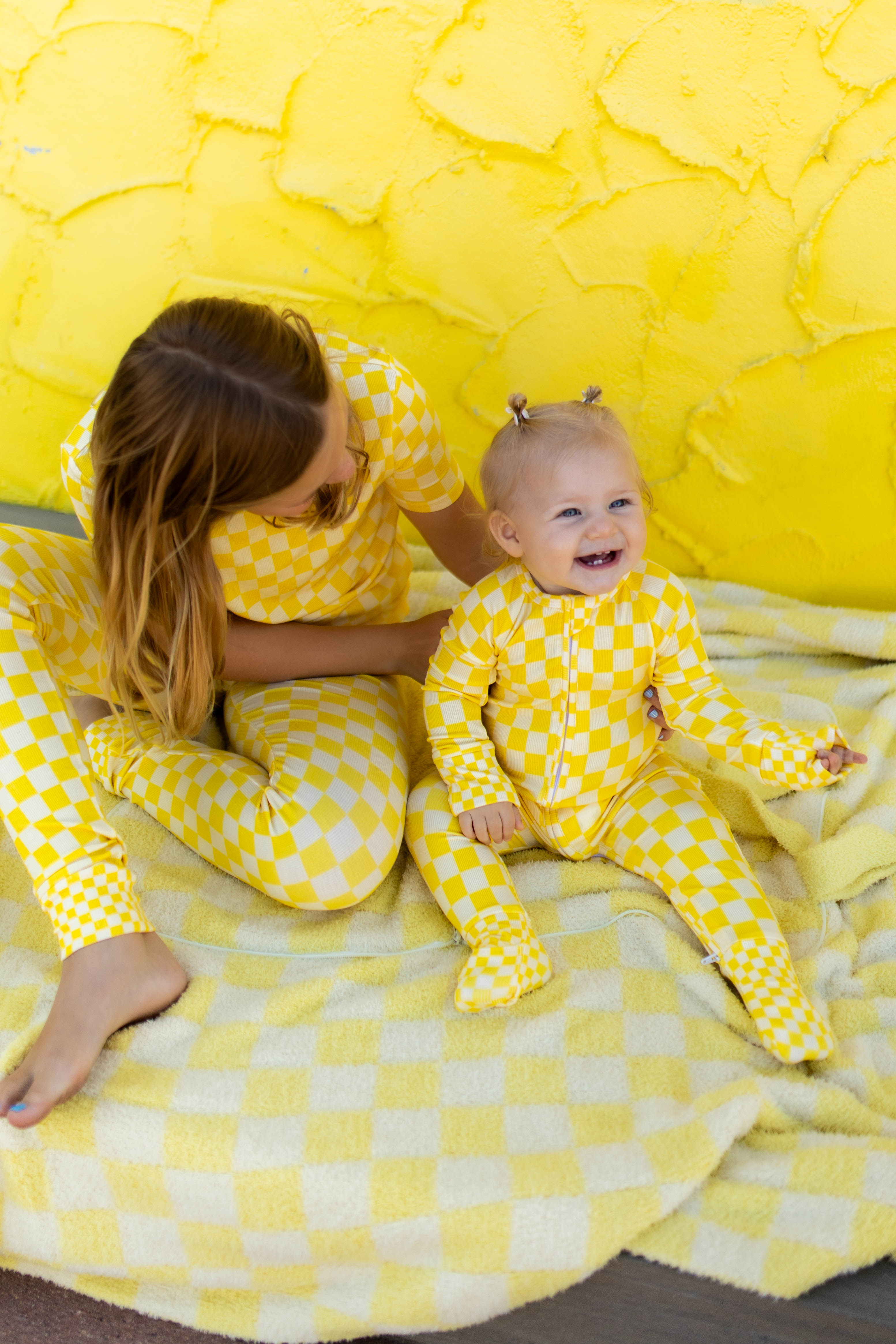 A girl and a baby wearing matching yellow and white checkered pajamas, sitting together on a yellow blanket in front of a textured yellow backdrop.