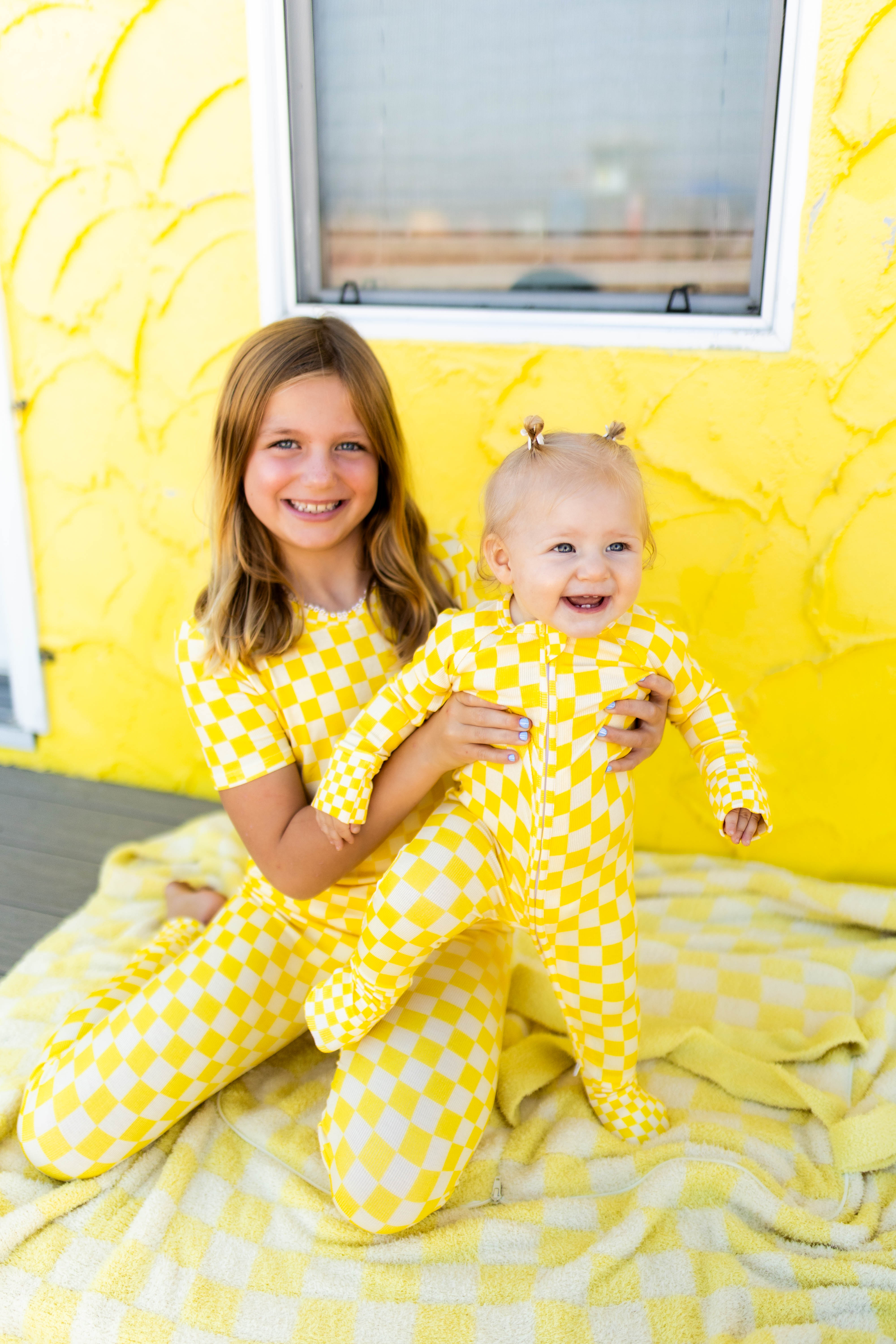 A smiling young girl and a baby sitting together on a yellow blanket in front of a textured yellow wall. Both are wearing matching yellow checkered footed pajamas.