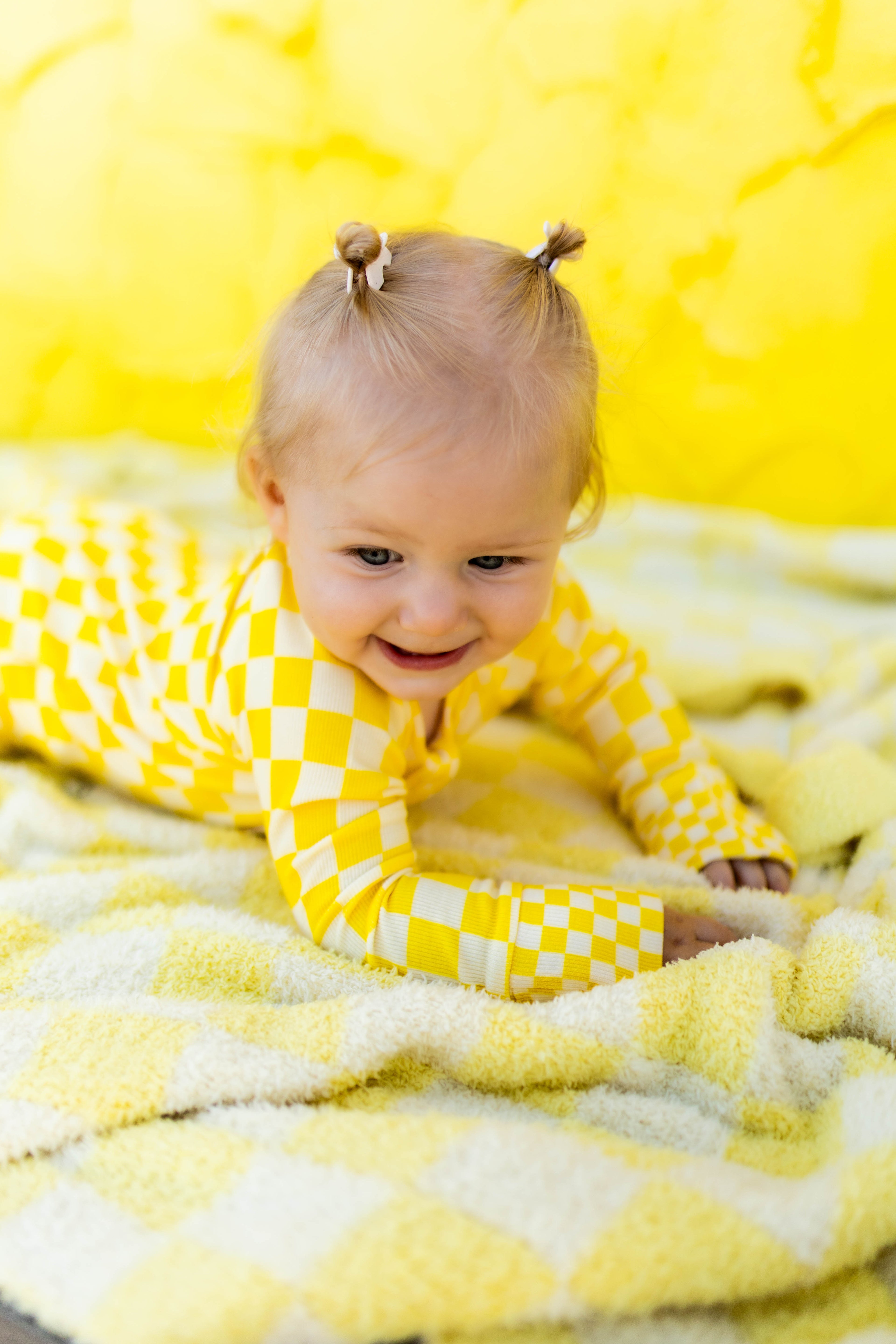 A baby girl wearing a yellow  checkered long-sleeve romper, crawling on a matching yellow blanket against a bright yellow background.
