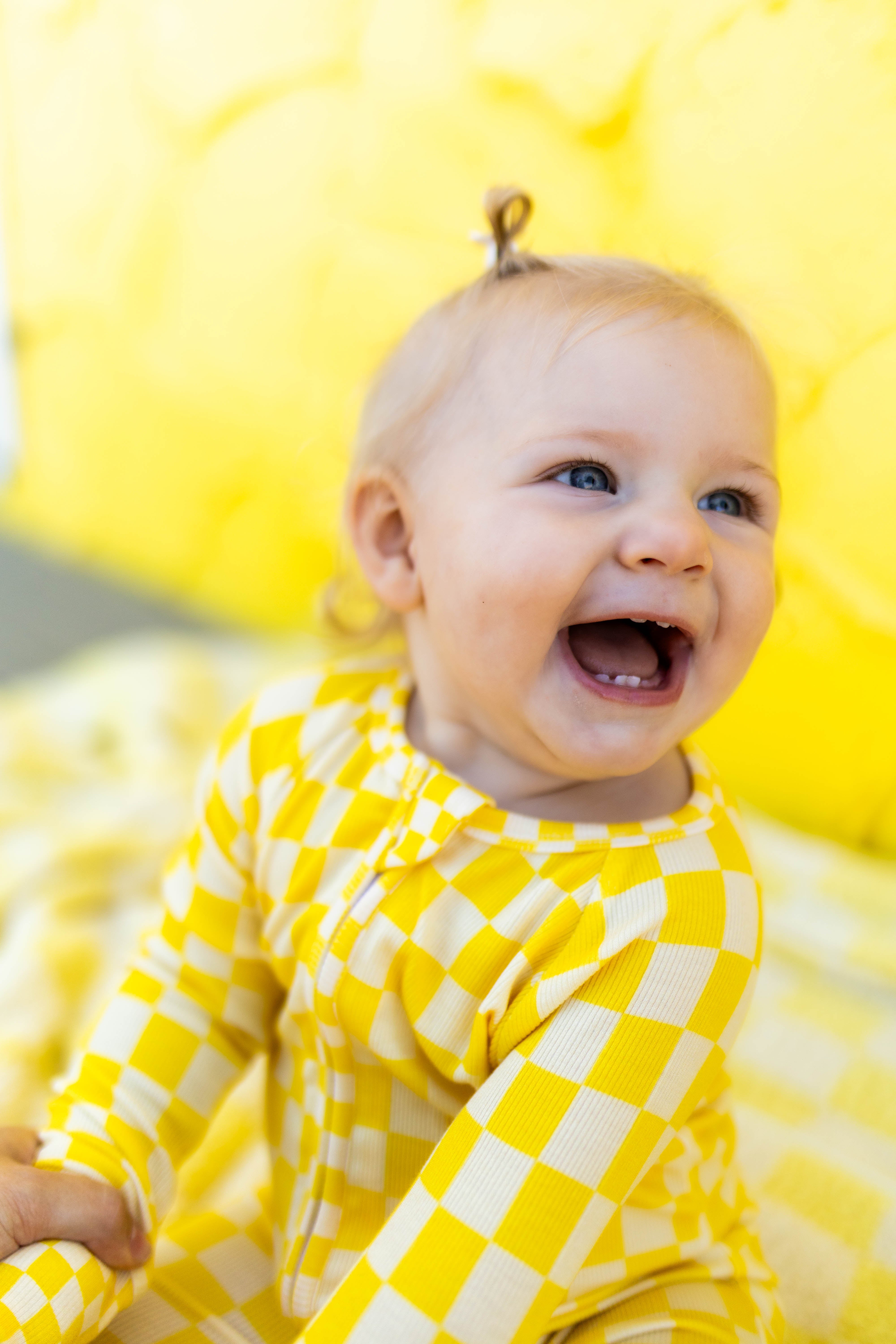 A baby wearing a yellow checkered long-sleeve romper, smiling and laughing while sitting against a bright yellow background.
