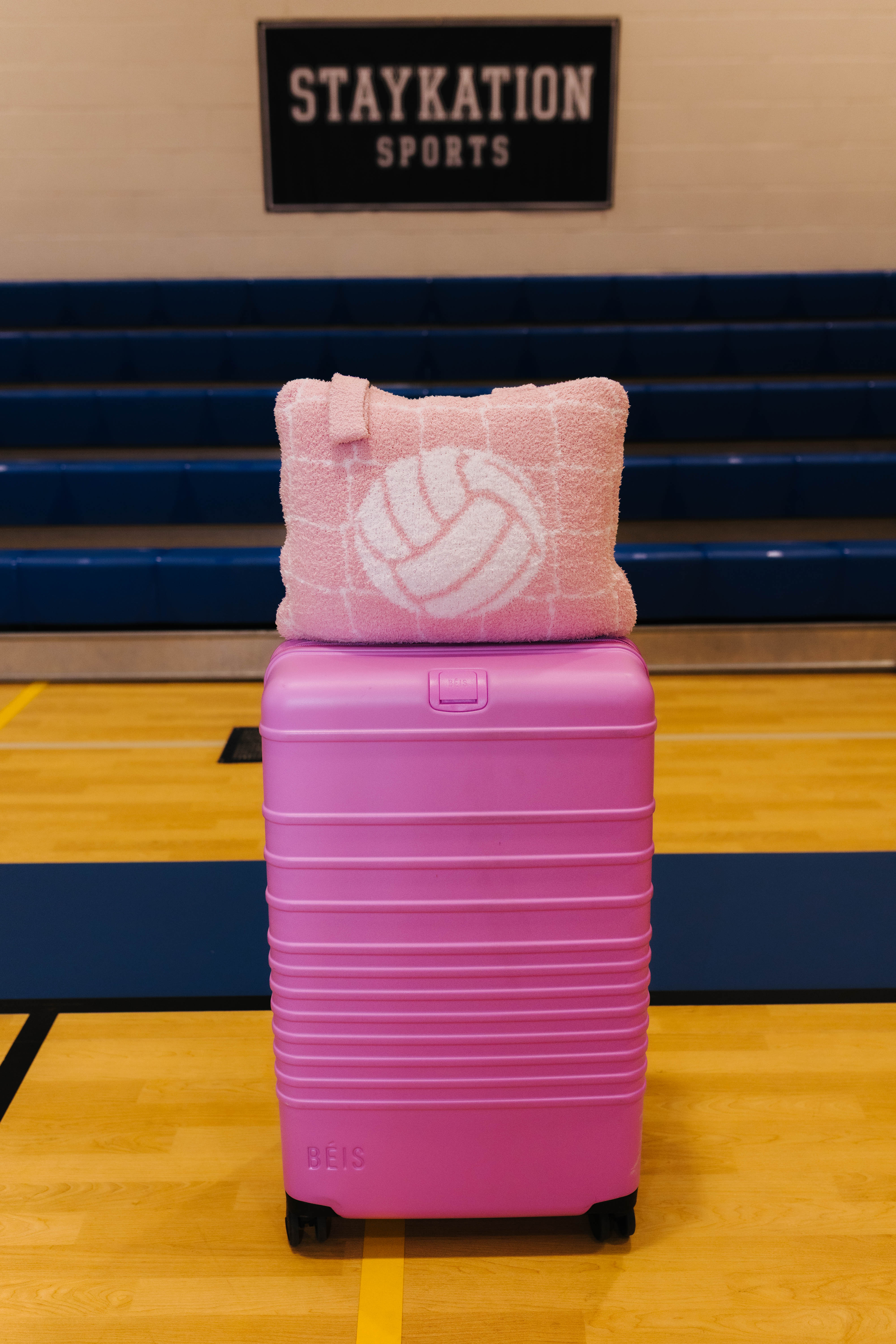 A pink hard-shell suitcase with black wheels sits on a gym floor in front of blue bleachers, with a pink quillow featuring a white volleyball pattern resting on top. A sign reading 'STAYKATION SPORTS' is visible in the background.