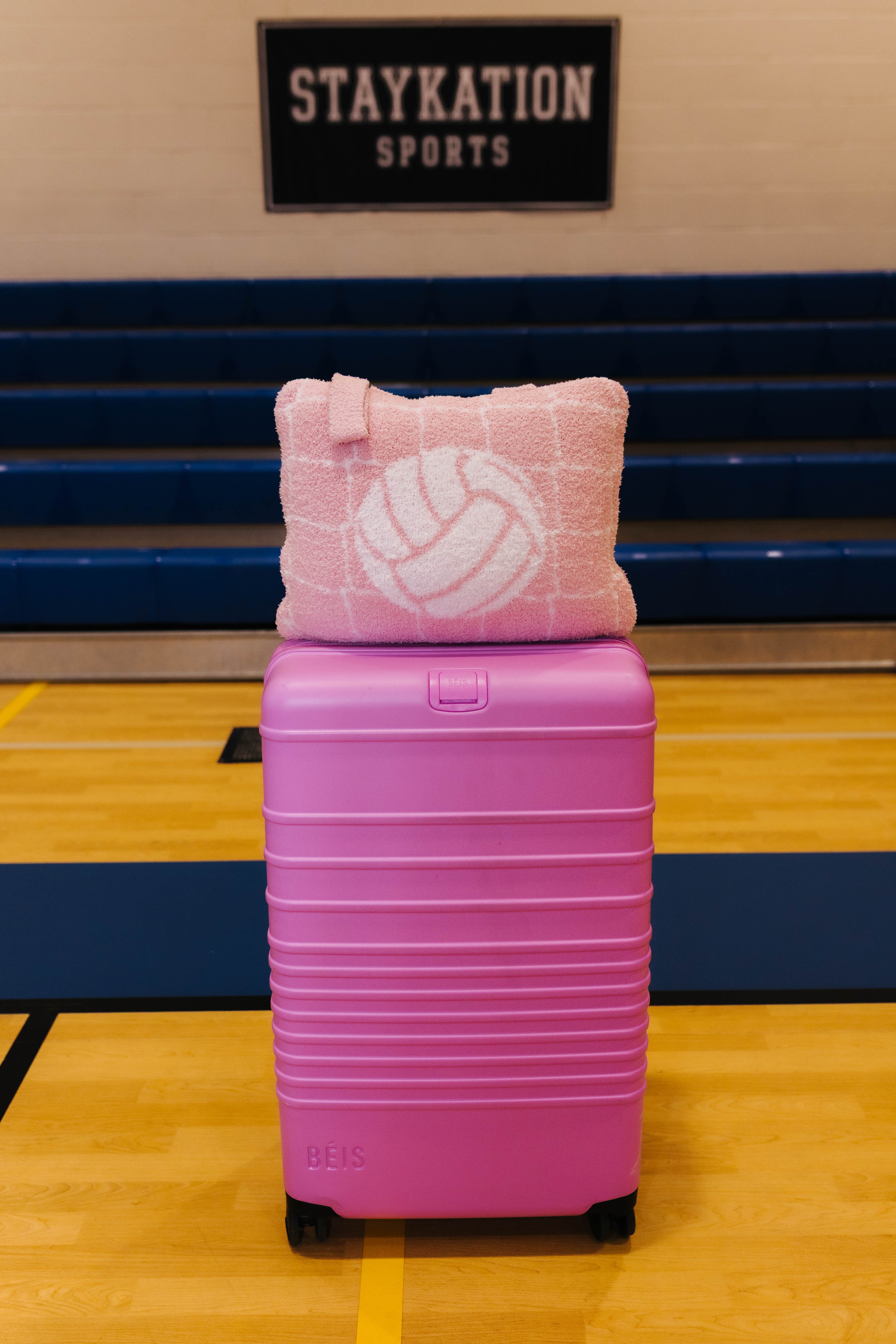 A pink hard-shell suitcase with black wheels sits on a gym floor in front of blue bleachers, with a pink quillow featuring a white volleyball pattern resting on top. A sign reading 'STAYKATION SPORTS' is visible in the background.