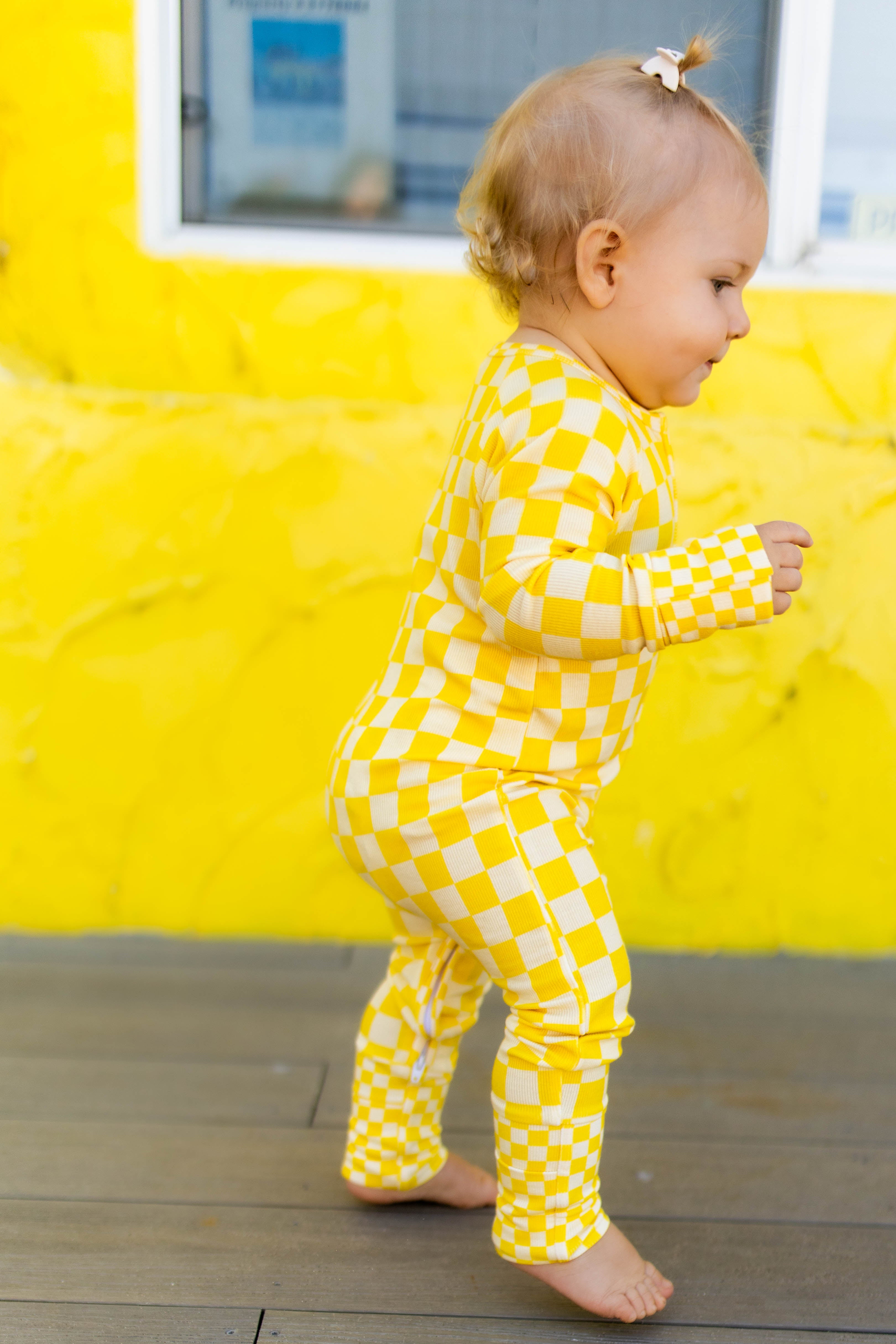 A toddler walking barefoot on a wooden deck in front of a bright yellow wall, wearing a yellow checkered long-sleeve romper.