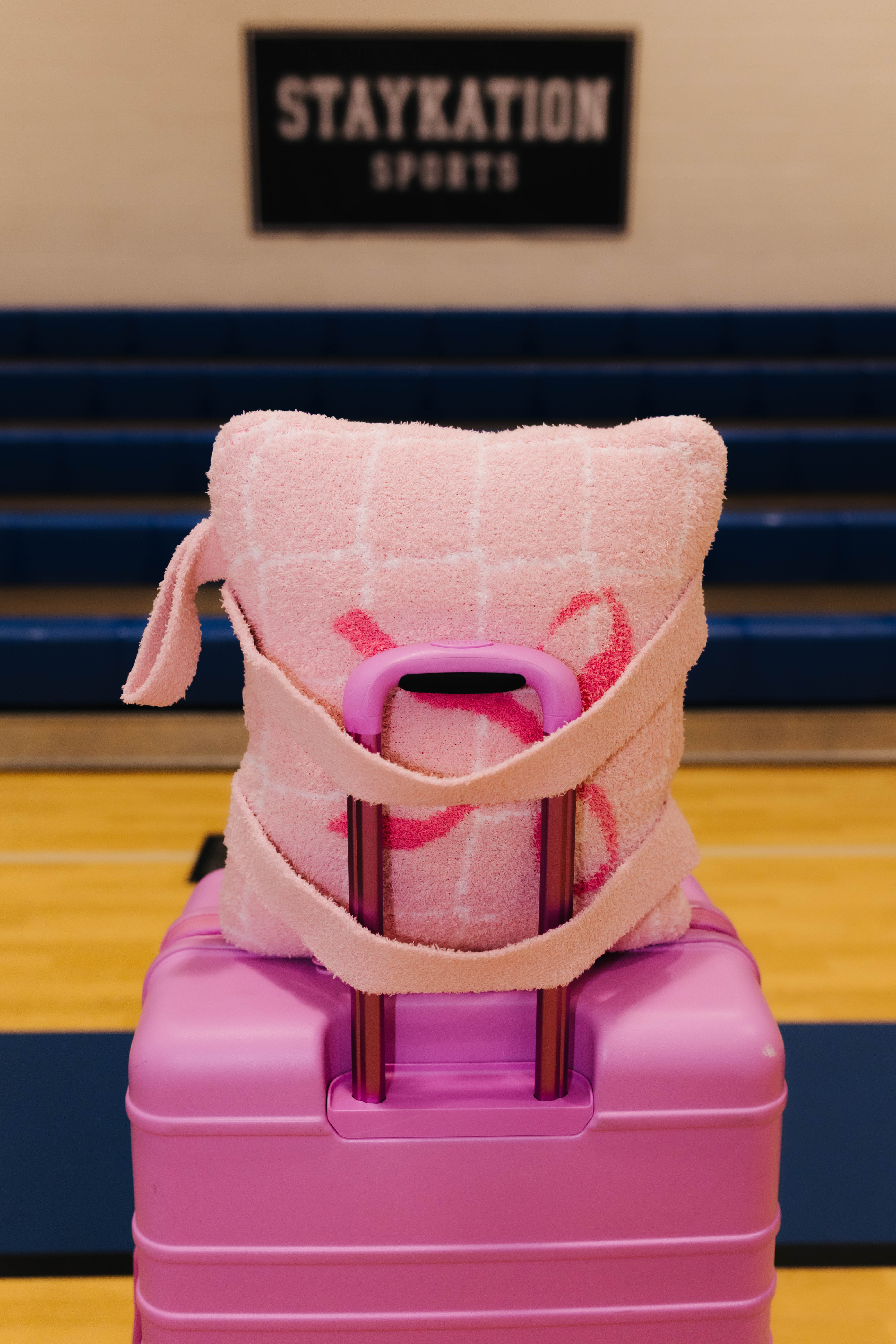 A pink quillow with a grid pattern and pink ribbon accents, wrapped in a matching pink towel strap, resting on top of a pink rolling suitcase in a gymnasium setting.