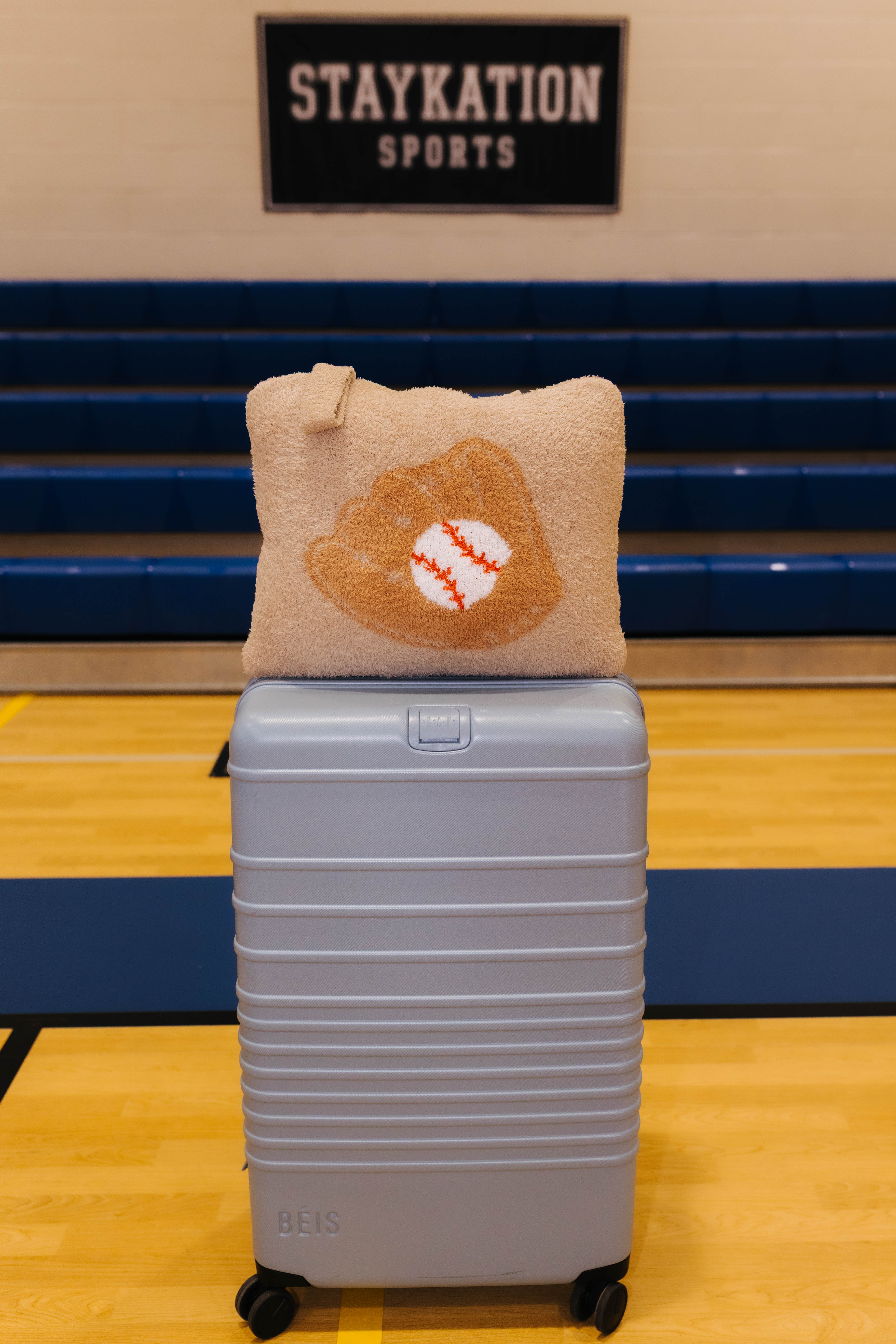 A light blue hard-shell rolling suitcase with a baseball quillow on top it has beige fabric and features a baseball glove design with a baseball in the center.