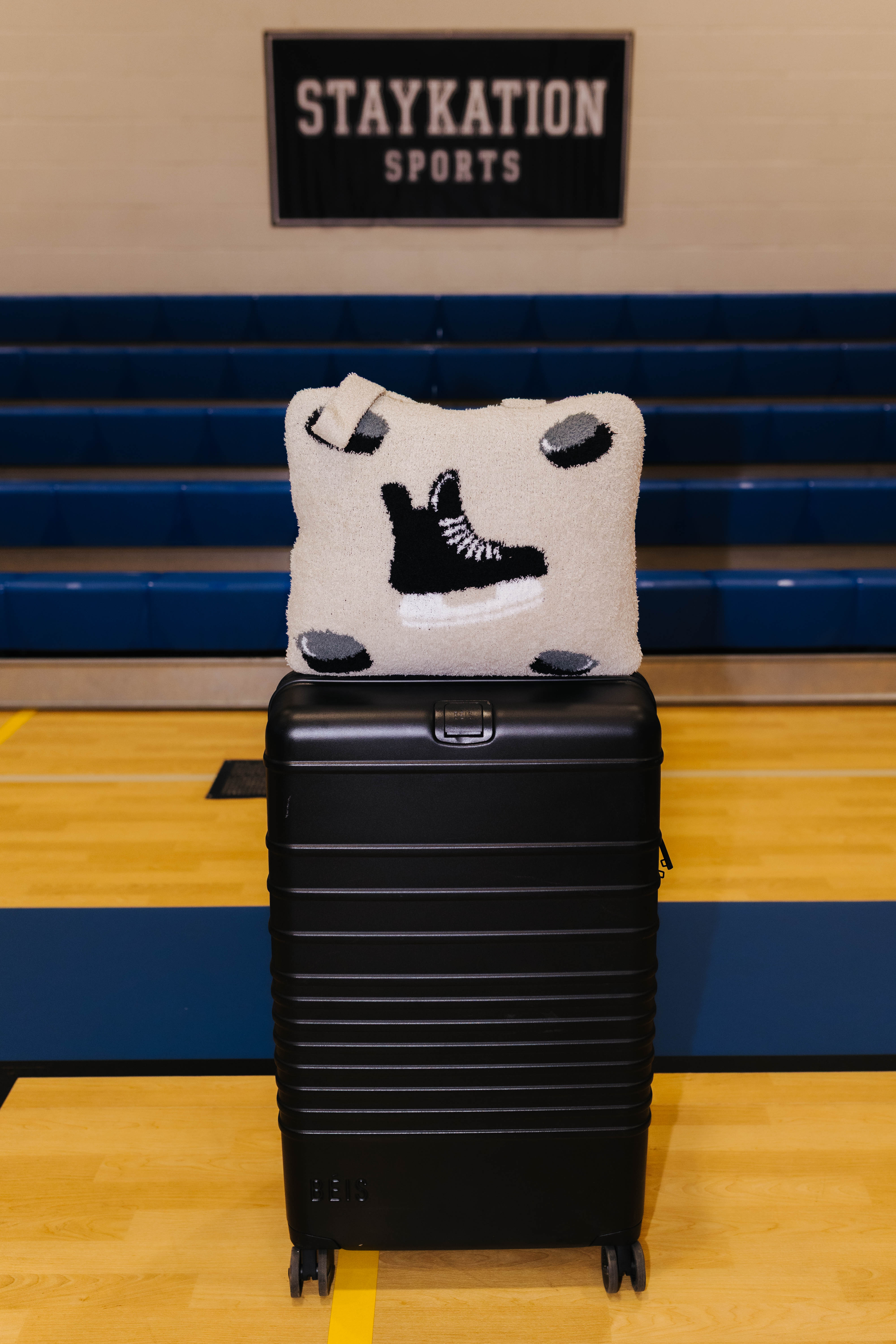 A black hard-shell suitcase standing on a wooden gym floor with blue bleachers and a wall sign in the background. On top of the suitcase is a quillow featuring a black hockey skate design.