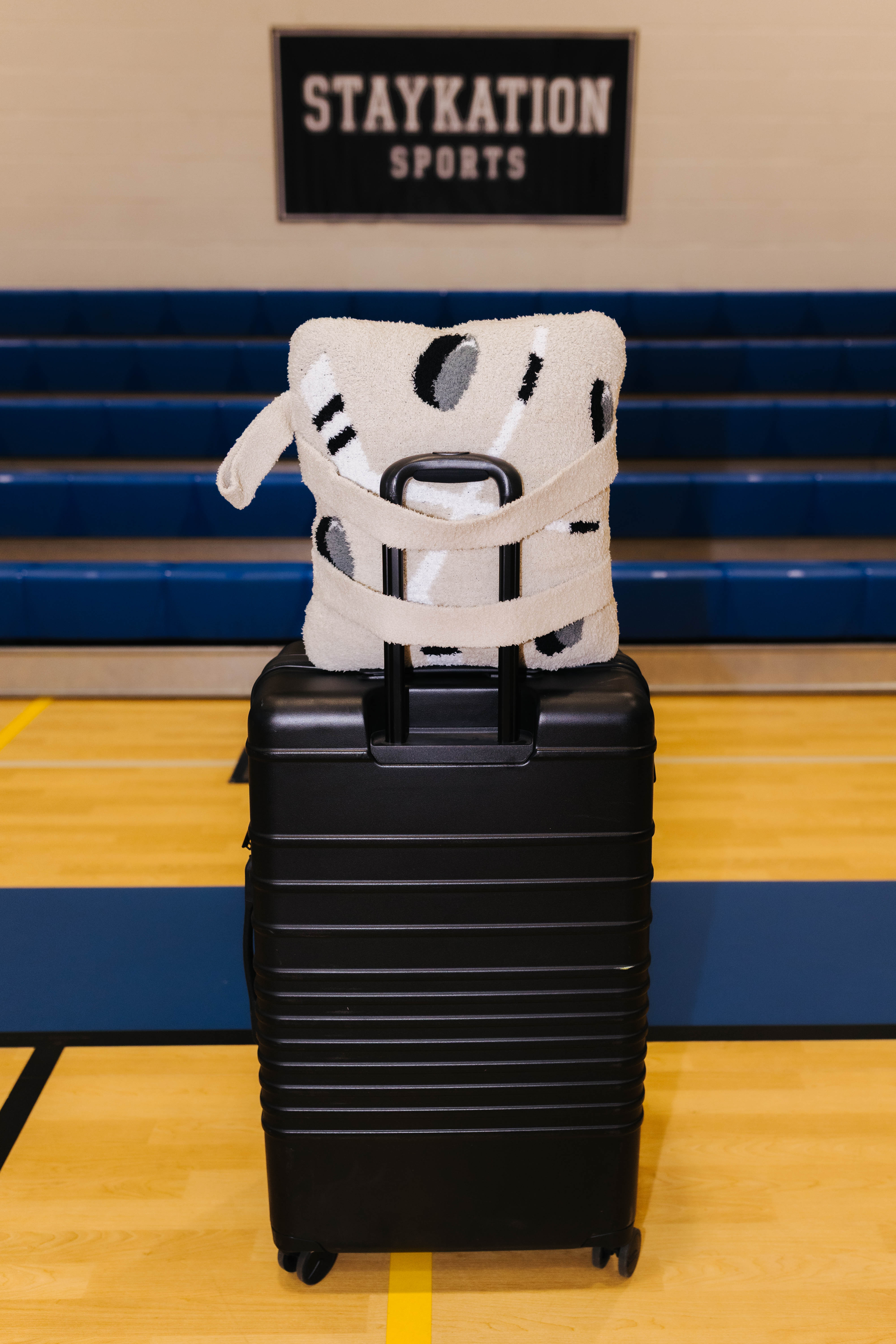 A black hard-shell rolling suitcase stands on a gym floor with a white and black patterned hockey quillow on top. Blue bleachers and a wall sign reading STAYKATION SPORTS are visible in the background.
