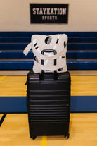 A black hard-shell rolling suitcase stands on a gym floor with a white and black patterned hockey quillow on top. Blue bleachers and a wall sign reading STAYKATION SPORTS are visible in the background.