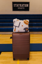 A brown hard-shell rolling suitcase stands on a gym floor in front of blue bleachers, with a tan quillow featuring football-themed graphics resting on top. A 'STAYKATION SPORTS' sign is visible on the wall behind.