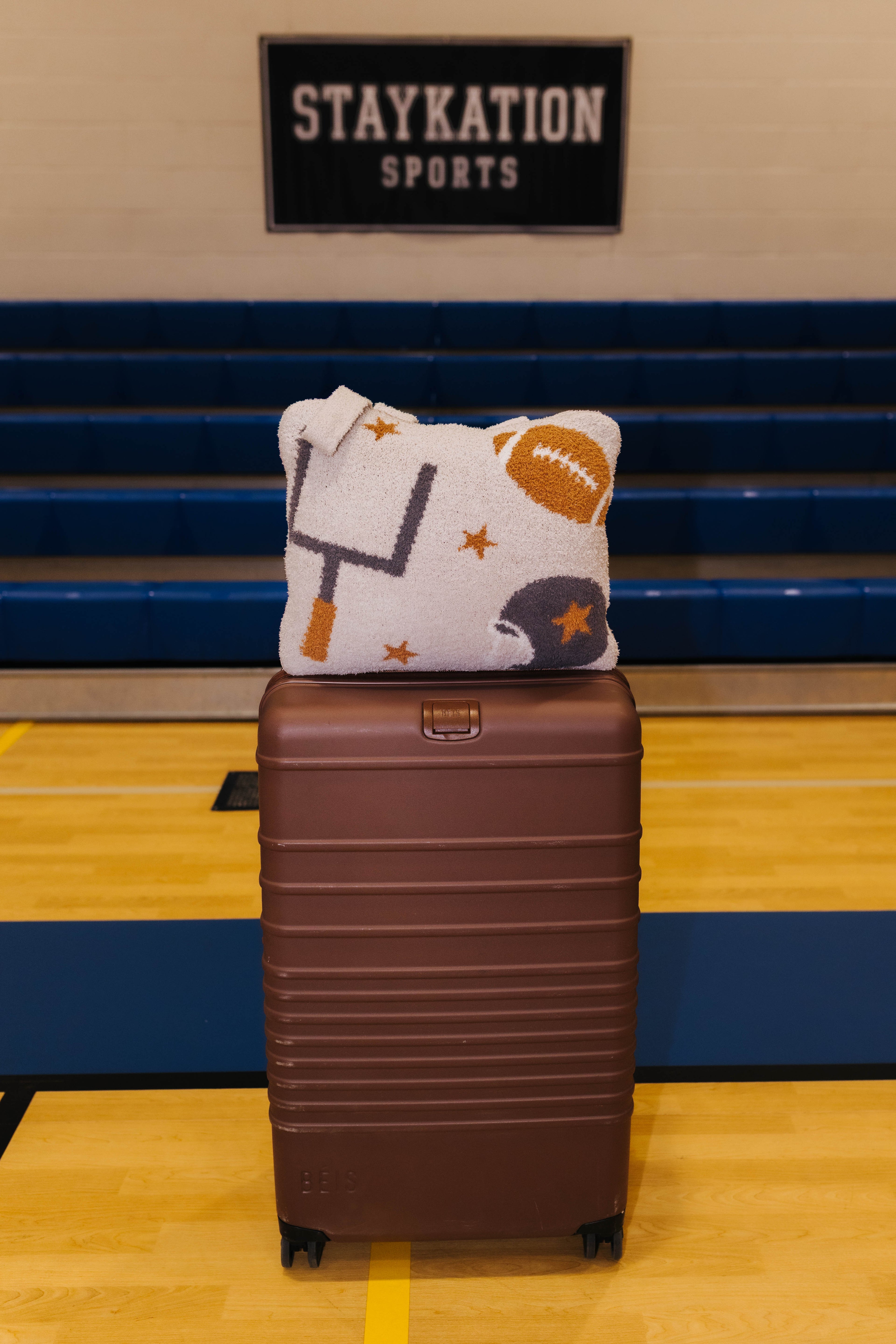 A brown hard-shell rolling suitcase stands on a gym floor in front of blue bleachers, with a tan quillow featuring football-themed graphics resting on top. A 'STAYKATION SPORTS' sign is visible on the wall behind.