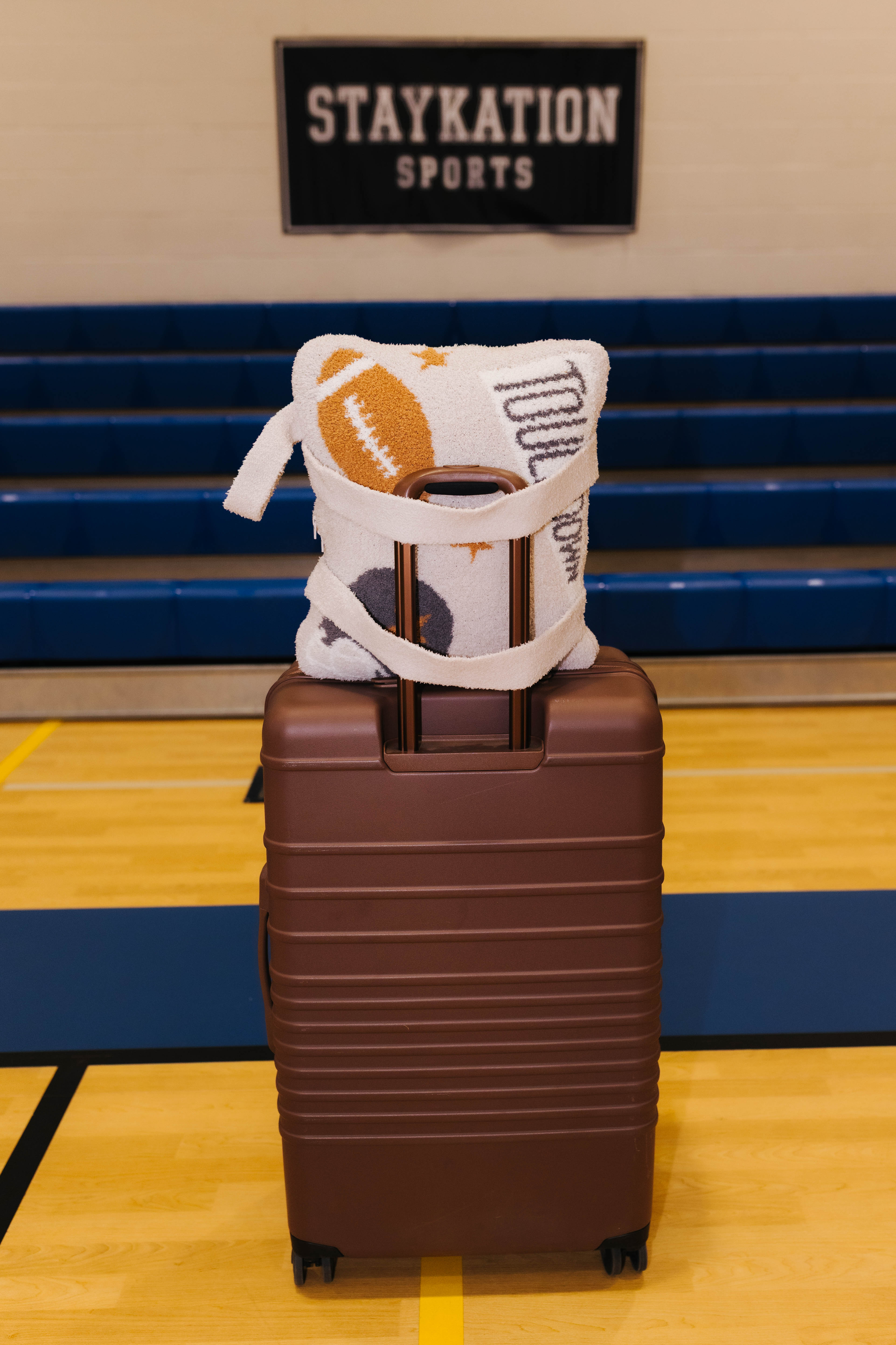 A brown hard-shell suitcase standing on a gym floor with the tan quillow with a football and printed with text, set against blue bleachers and a sign reading STAYKATION SPORTS.