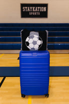 A bright blue hard-shell rolling suitcase stands on a gym floor in front of blue bleachers, with a black and gray knit quillow featuring a soccer ball design resting on top. 