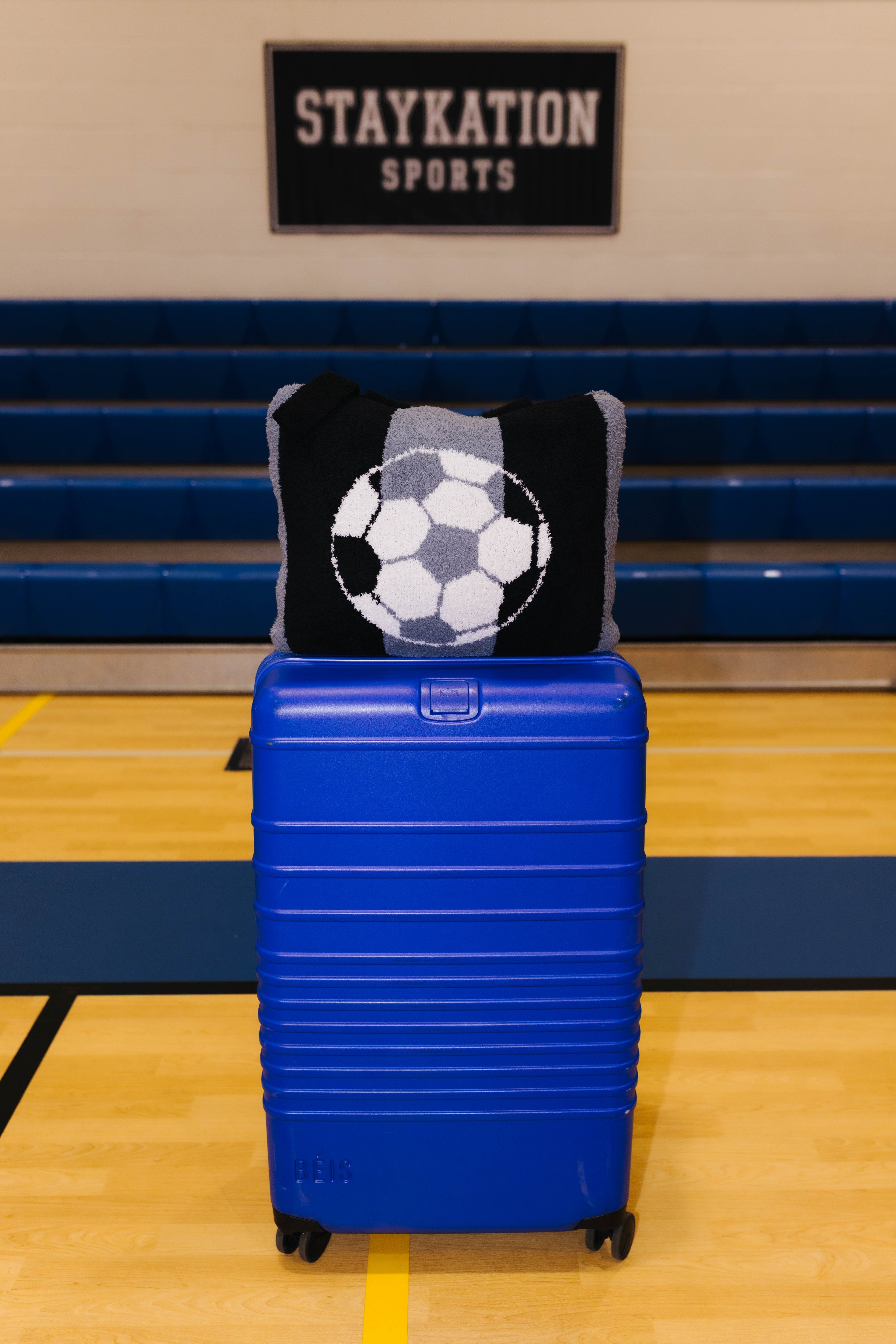 A bright blue hard-shell rolling suitcase stands on a gym floor in front of blue bleachers, with a black and gray knit quillow featuring a soccer ball design resting on top. 