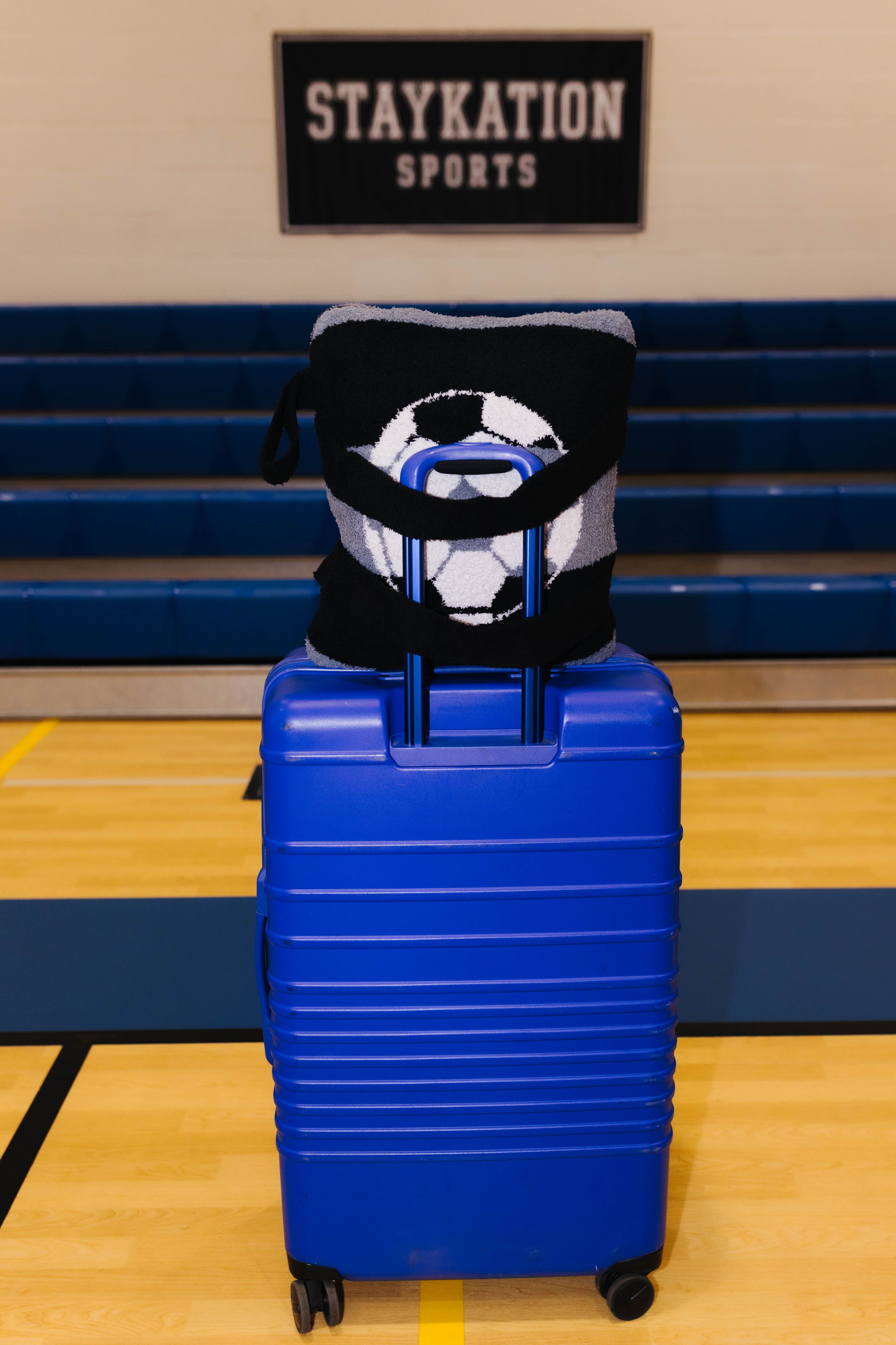 A bright blue hard-shell rolling suitcase stands on an indoor gym floor showing the backside of the quillow.