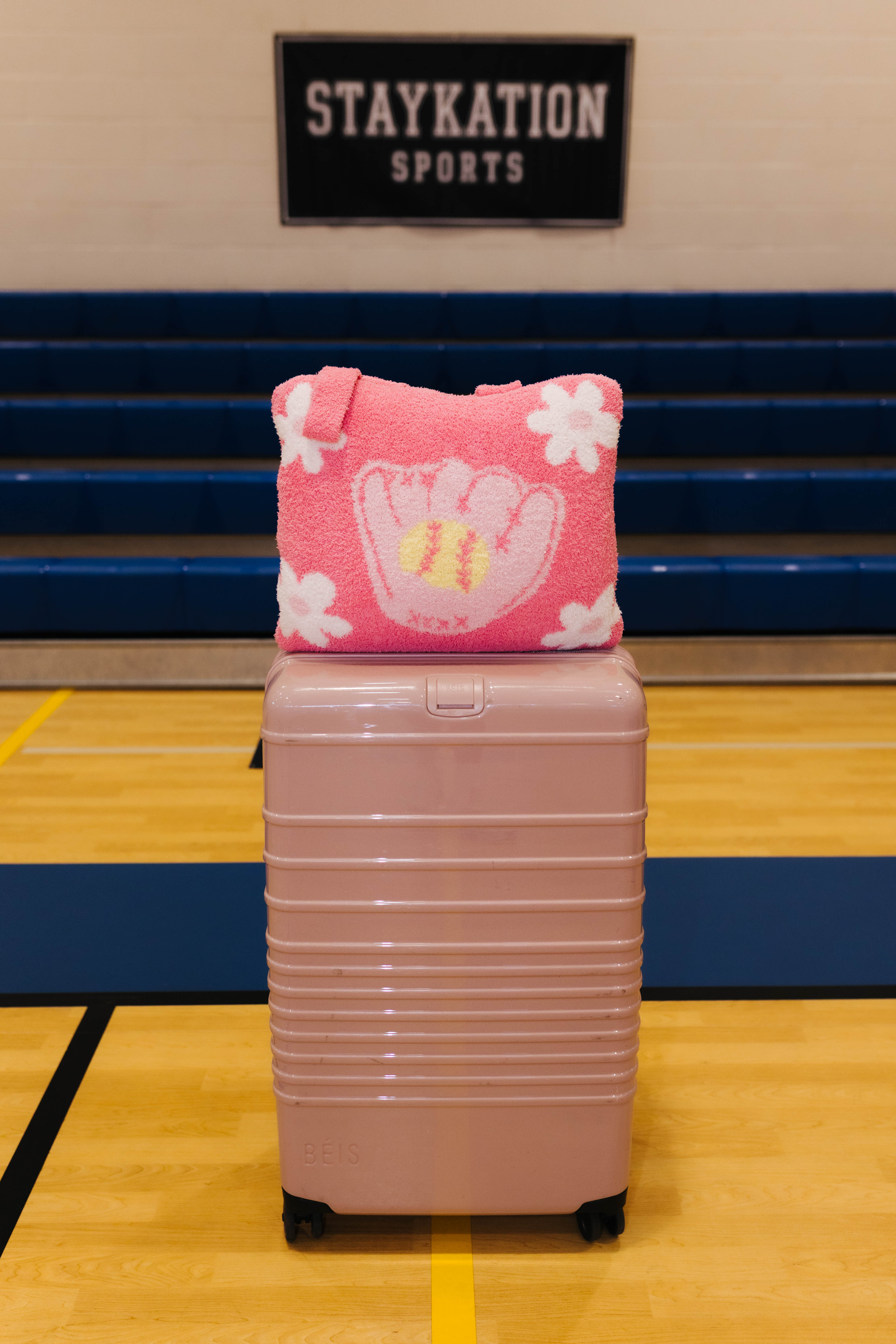 A pink quillow white baseball glove and flower pattern sits on top of a dusty pink hard-shell suitcase in a gymnasium. A 'STAYKATION SPORTS' sign is visible in the background.