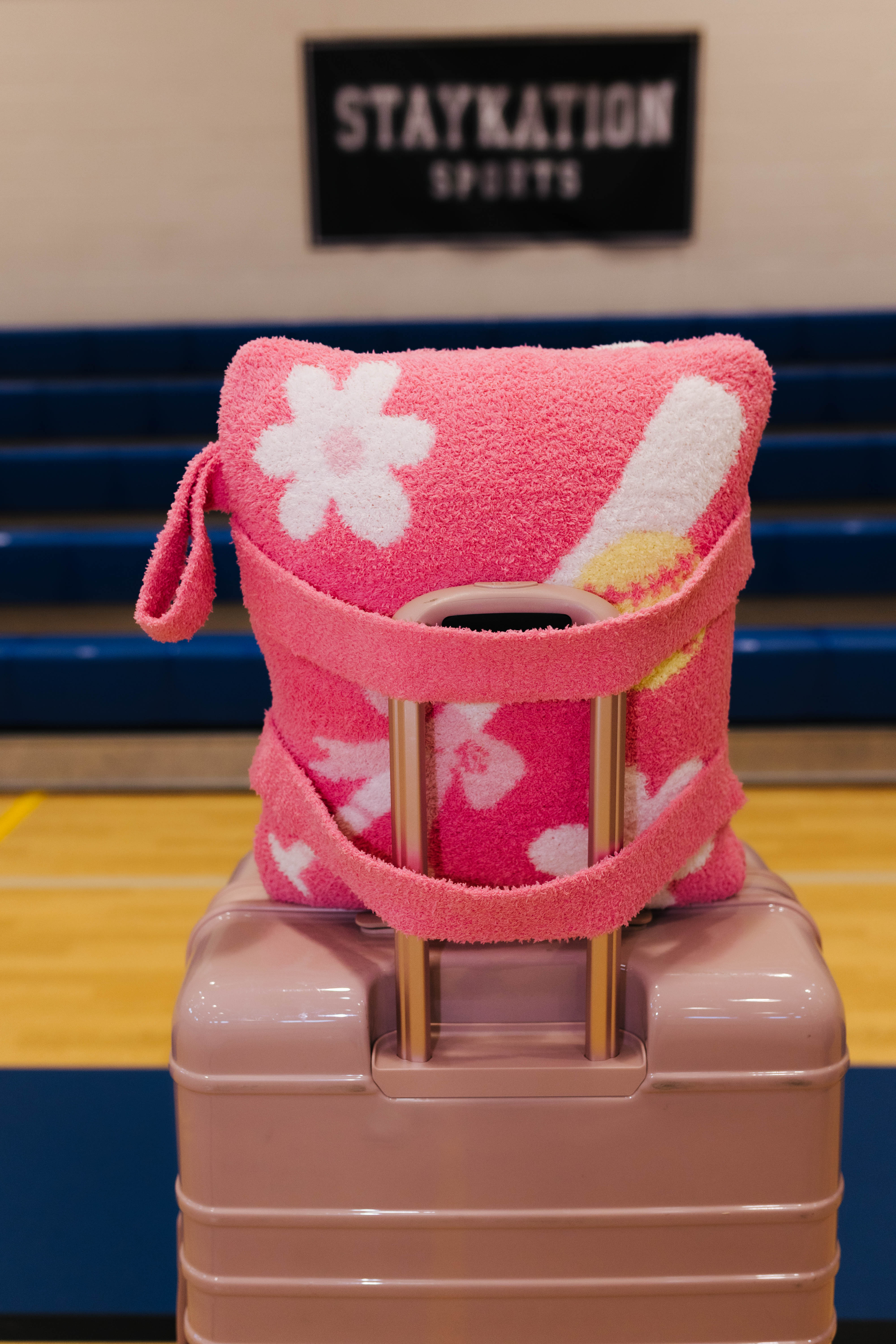 A pink quillow white floral patterns and a pink strap, wrapped around a light pink suitcase in a gym setting with a 'STAYKATION SPORTS' sign in the background.
