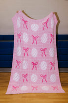 A blanket featuring a repeating pattern of white volleyballs and pink bows, hanging against a wall with blue lockers in the background.