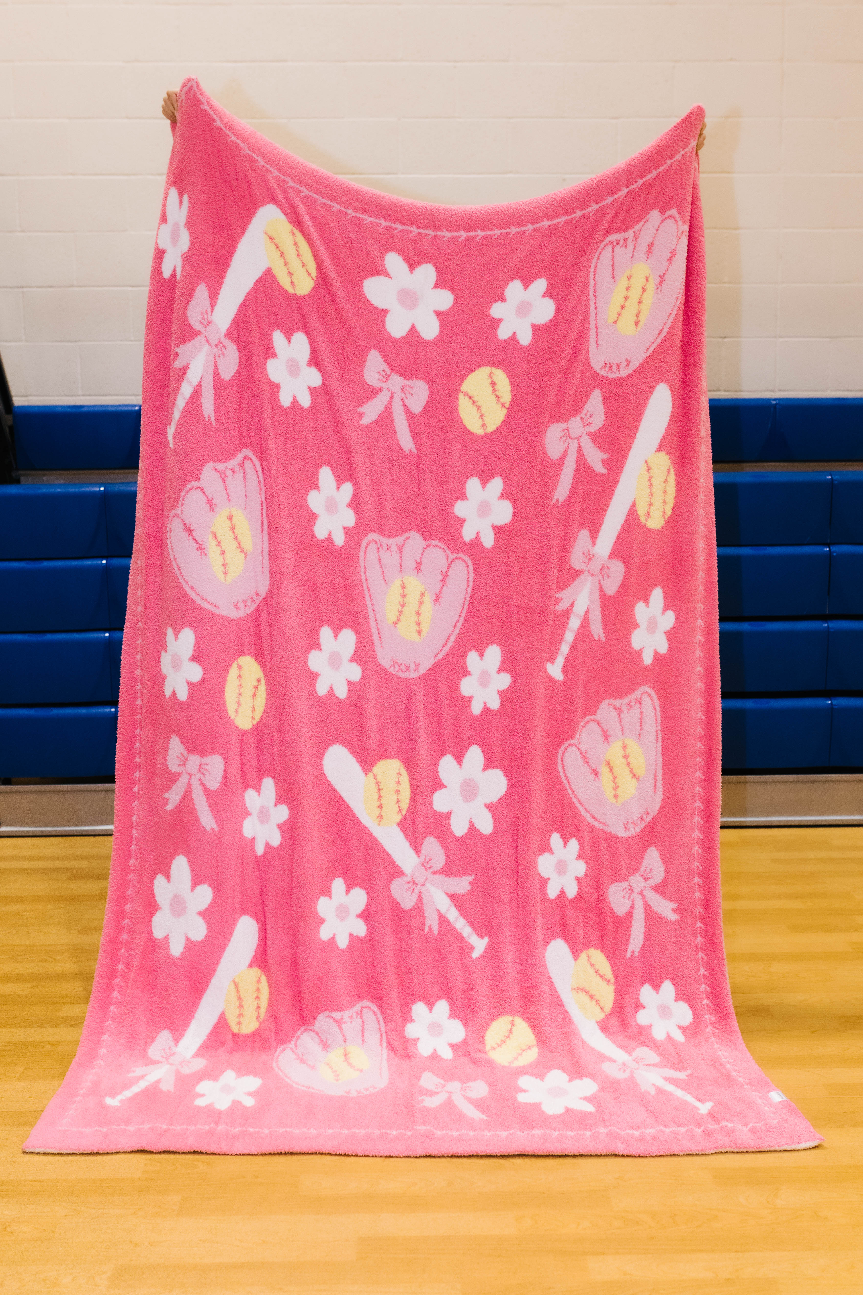 A pink fleece blanket with a baseball and softball theme, featuring white flowers, pink baseball gloves, yellow softballs, and white bats, hanging on a gym wall.