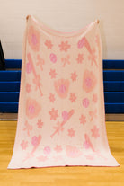 Back of the softball blanket showing light pink knit blanket with a whimsical pattern of pink flowers, bows, and hearts, hanging from a wooden rod in a gymnasium with blue bleachers in the background.