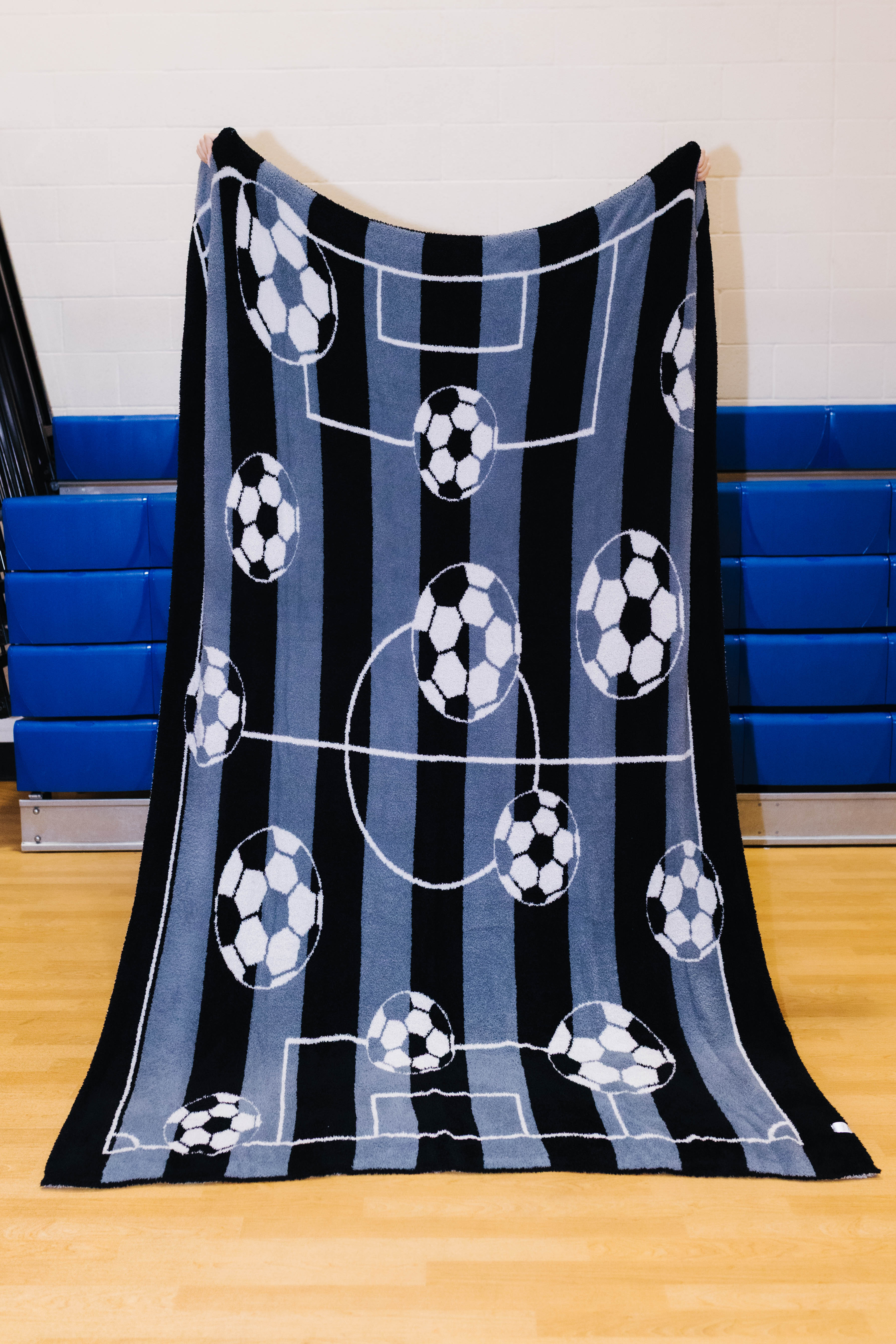A fleece throw blanket featuring a soccer theme with black and blue vertical stripes, white field lines, and soccer balls, held up by a person in an indoor gym setting.