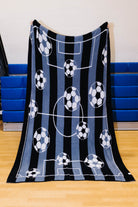 A fleece throw blanket featuring a soccer theme with black and blue vertical stripes, white field lines, and soccer balls, held up by a person in an indoor gym setting.