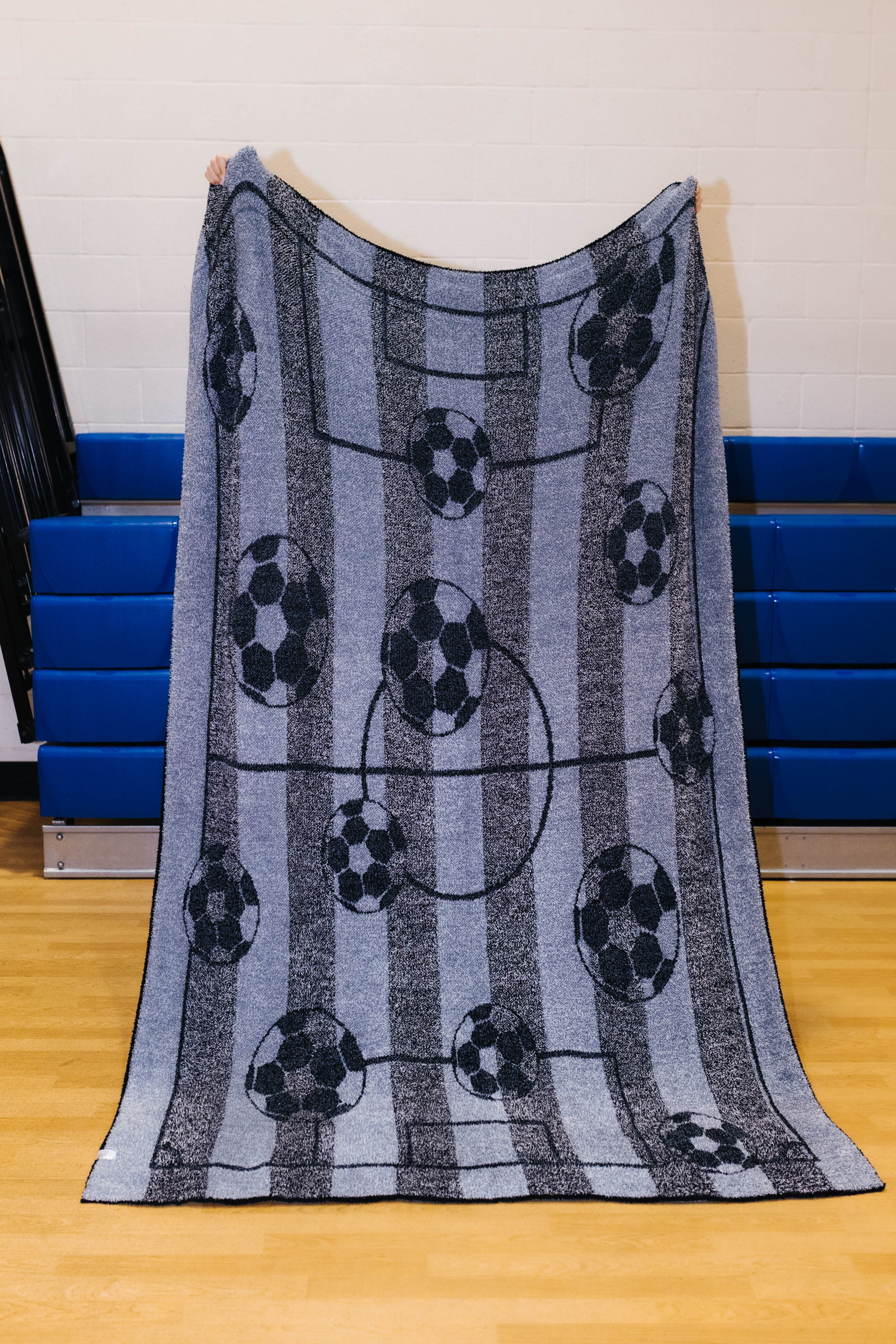 Back side of the rectangular fleece blanket with a soccer theme, featuring black and white soccer balls on vertical gray and blue-gray stripes with a faint soccer field graphic, held up by a person in an indoor gym setting.