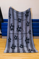 Back side of the rectangular fleece blanket with a soccer theme, featuring black and white soccer balls on vertical gray and blue-gray stripes with a faint soccer field graphic, held up by a person in an indoor gym setting.