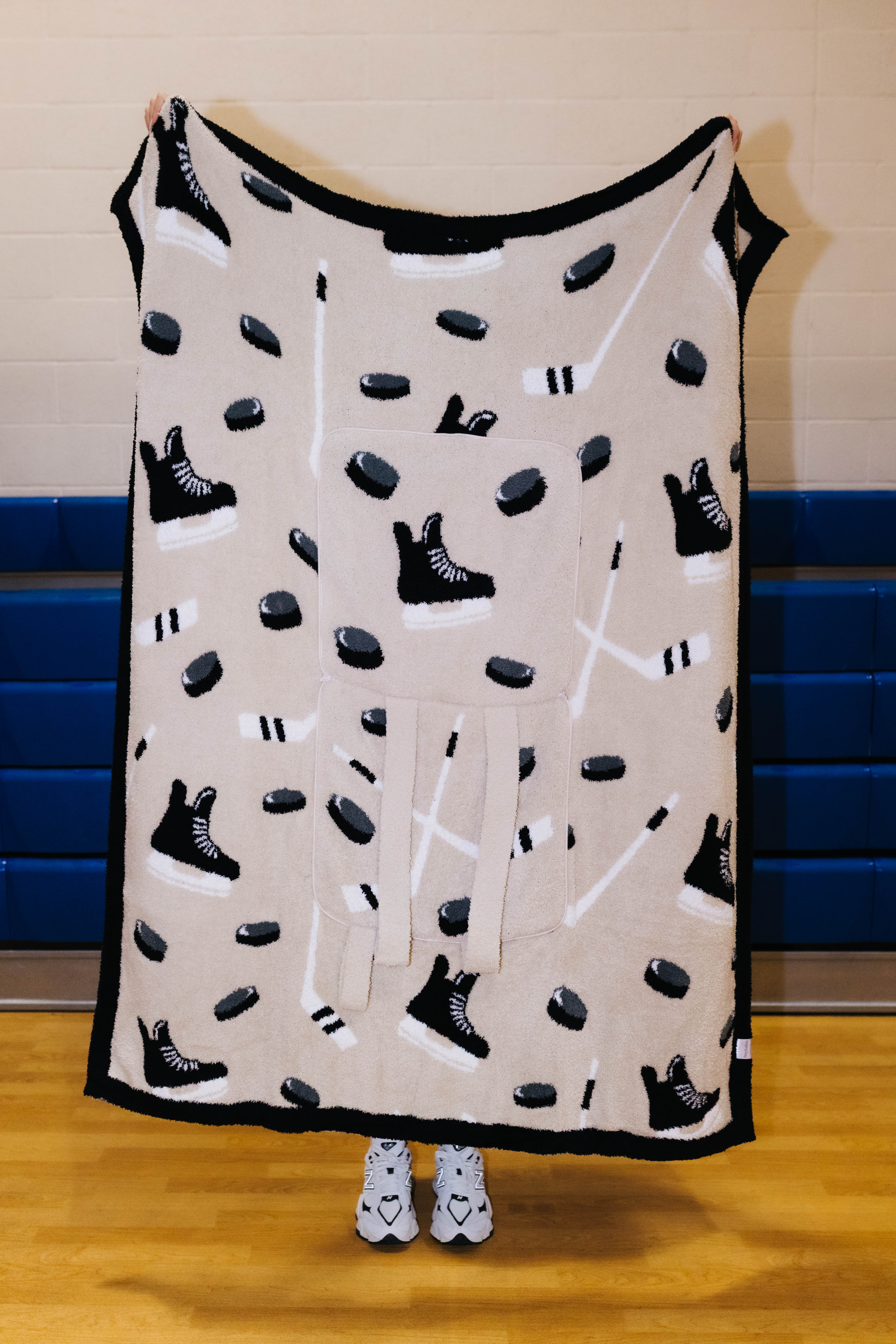 A fleece blanket with a hockey theme, featuring black skates, pucks, and sticks on a beige background with a black border, held up by a person in a gym.