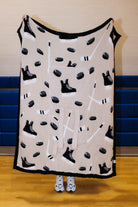 A fleece blanket with a hockey theme, featuring black skates, pucks, and sticks on a beige background with a black border, held up by a person in a gym.