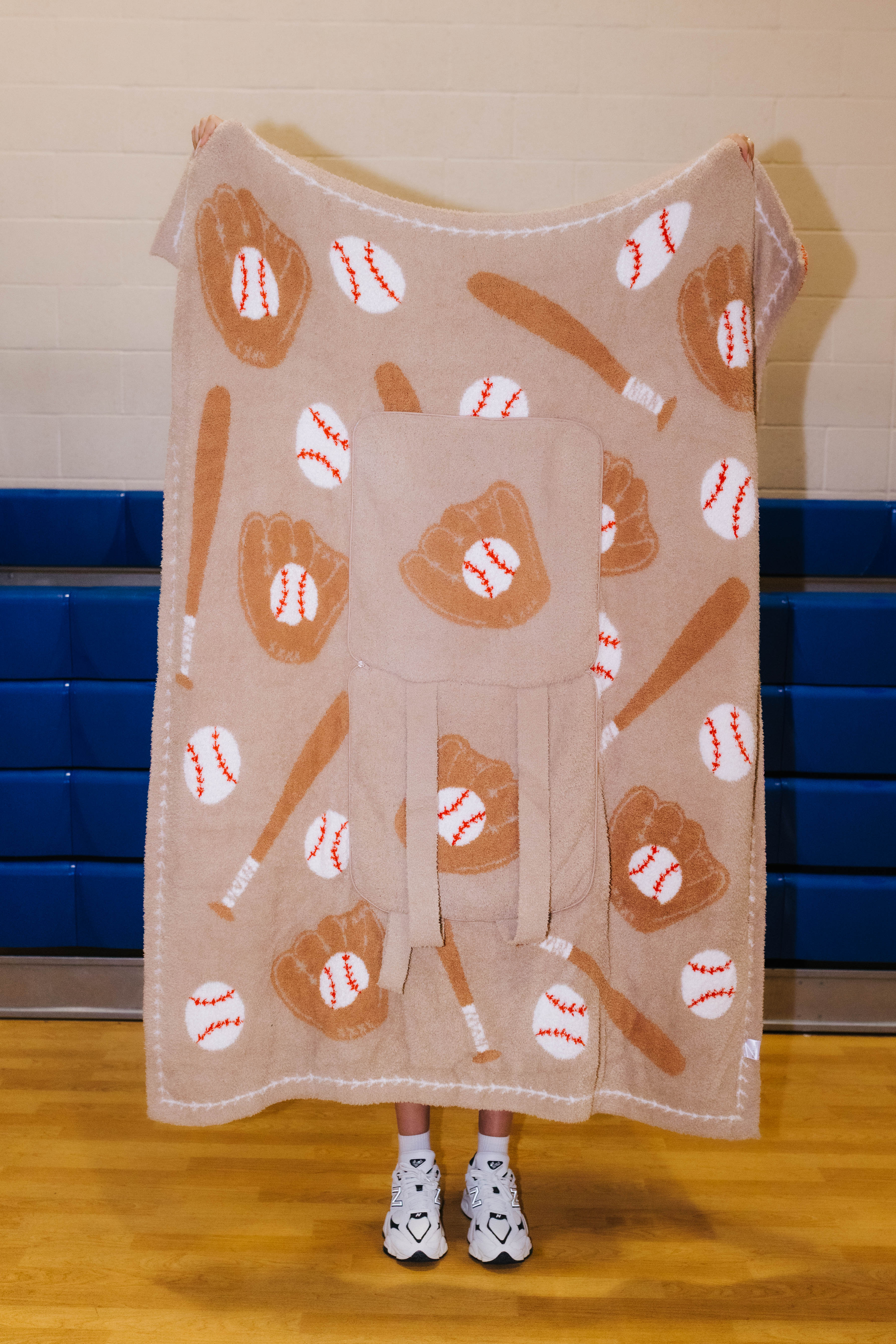 A person stands in a gym holding up a large fleece blanket or throw with a baseball theme. The blanket features an all-over print of brown baseball gloves, brown bats, and white baseballs with red stitching on a light tan background.