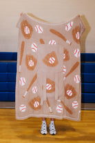 A person stands in a gym holding up a large fleece blanket or throw with a baseball theme. The blanket features an all-over print of brown baseball gloves, brown bats, and white baseballs with red stitching on a light tan background.