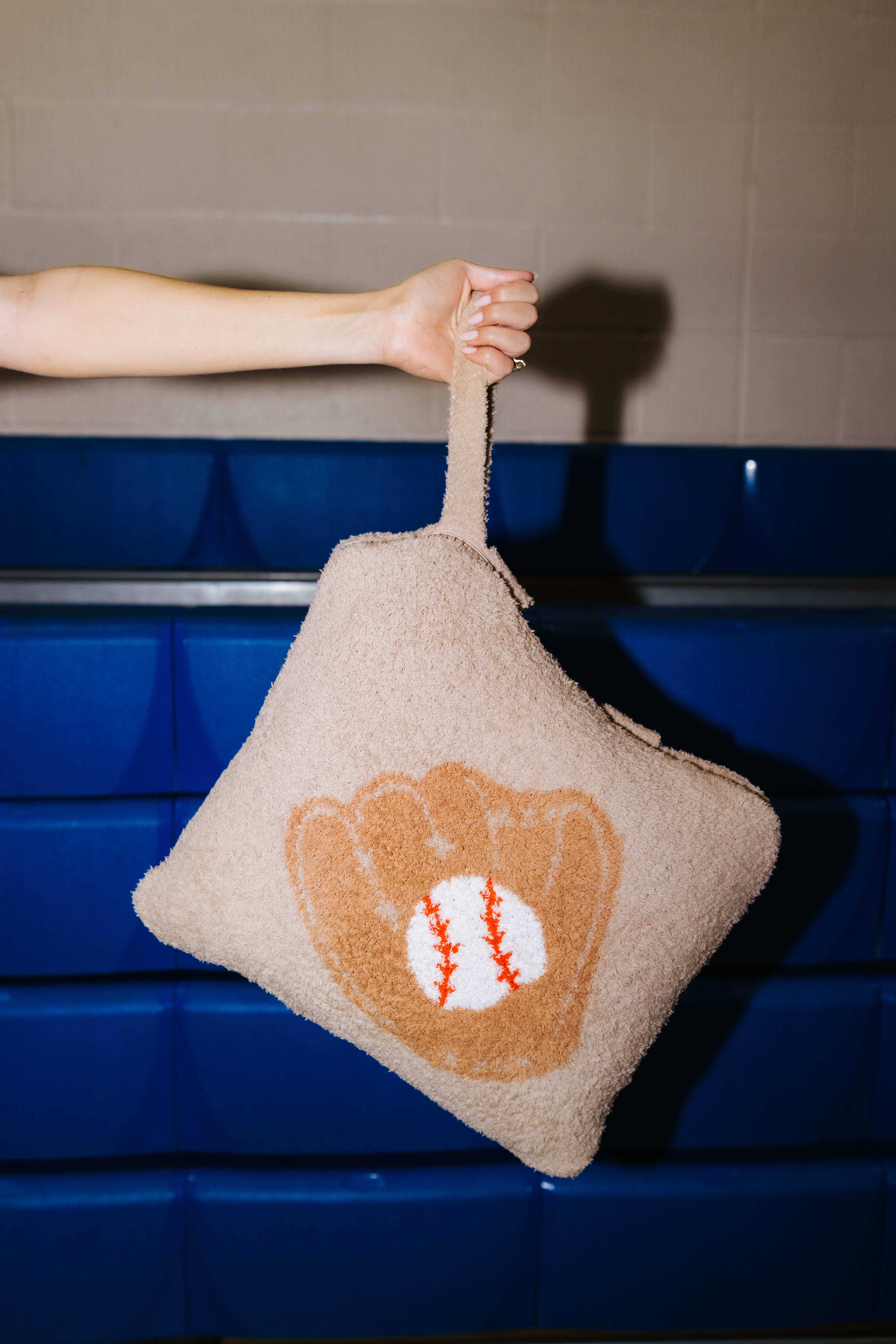 A beige baseball-themed quillow held by a person's arm against a blue padded wall and gray tiled background.