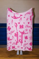 A pink fleece blanket with a pink and white cheerleader-themed print featuring dancers, bows, lipstick tubes, stars, and the word 'Cheer', held up by a person standing in front of a gym wall.