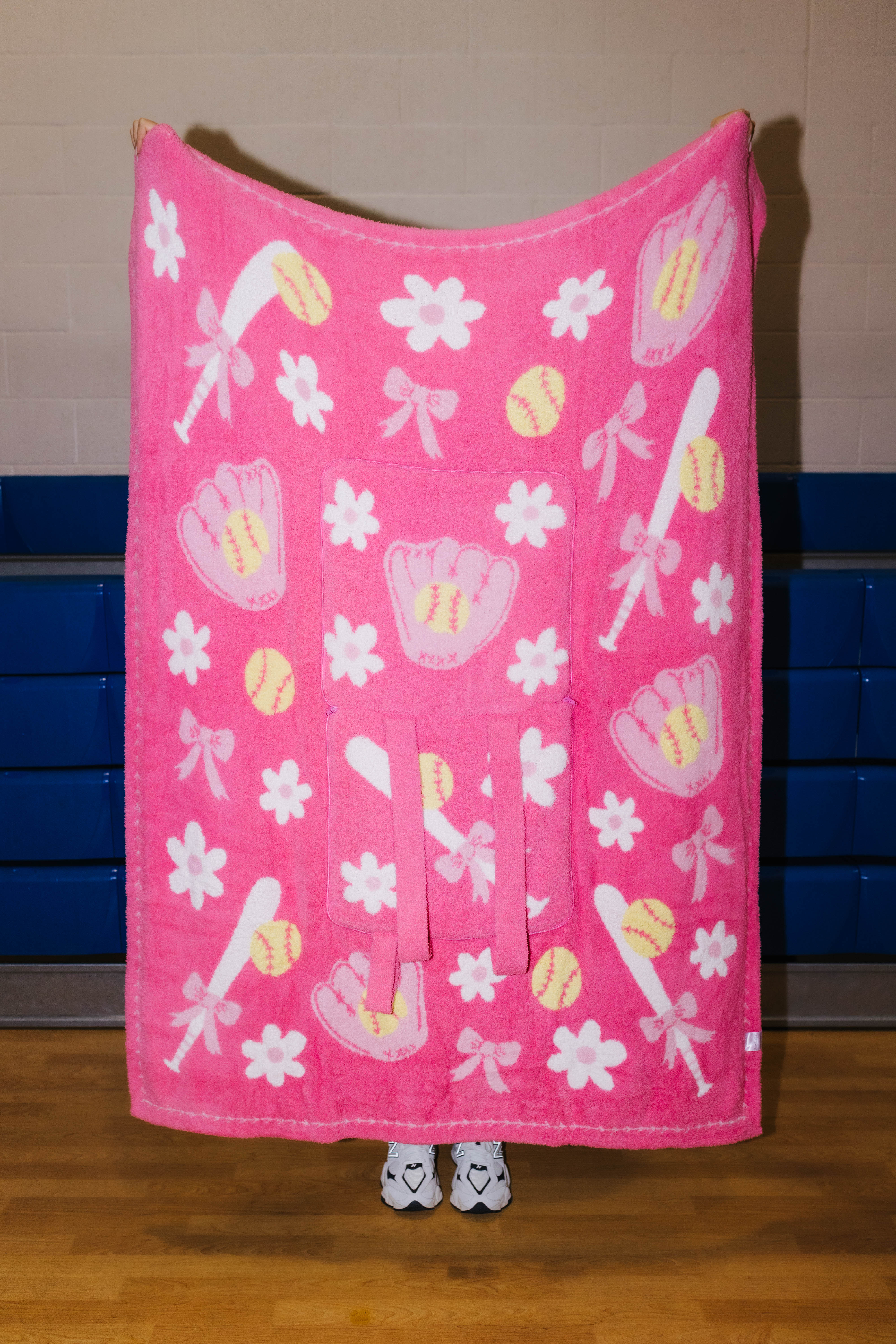 A pink fleece blanket with a baseball and softball pattern, held up by a person whose feet are visible at the bottom. The blanket features white flowers, pink bows, pink baseball gloves, and yellow baseballs and softballs.
