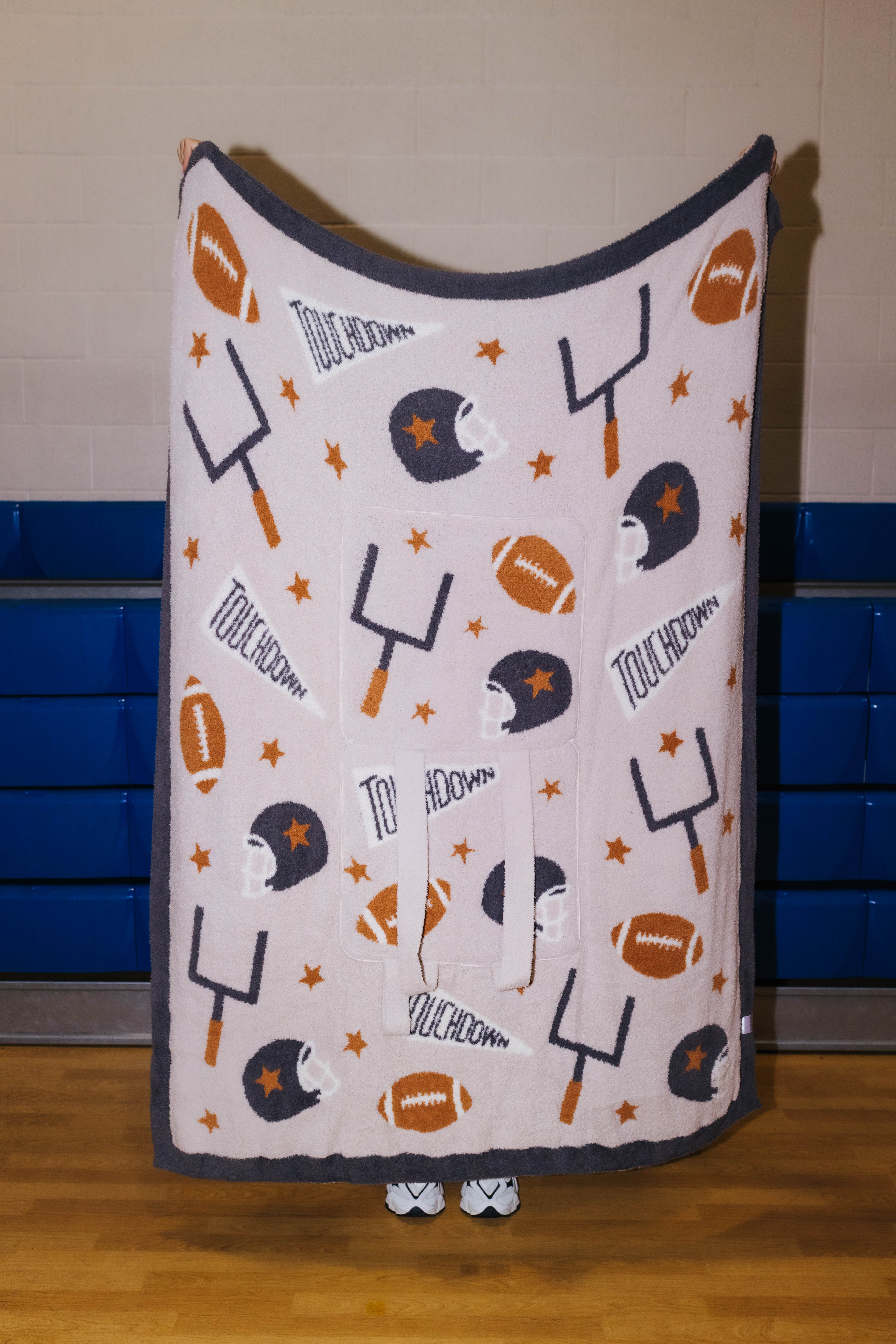 A fleece throw blanket with a football theme, featuring footballs, helmets, yardsticks, stars, and touchdown text in dark gray, orange, and white, held up by a person in a gymnasium.