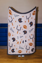 A fleece throw blanket with a football theme, featuring footballs, helmets, yardsticks, stars, and touchdown text in dark gray, orange, and white, held up by a person in a gymnasium.