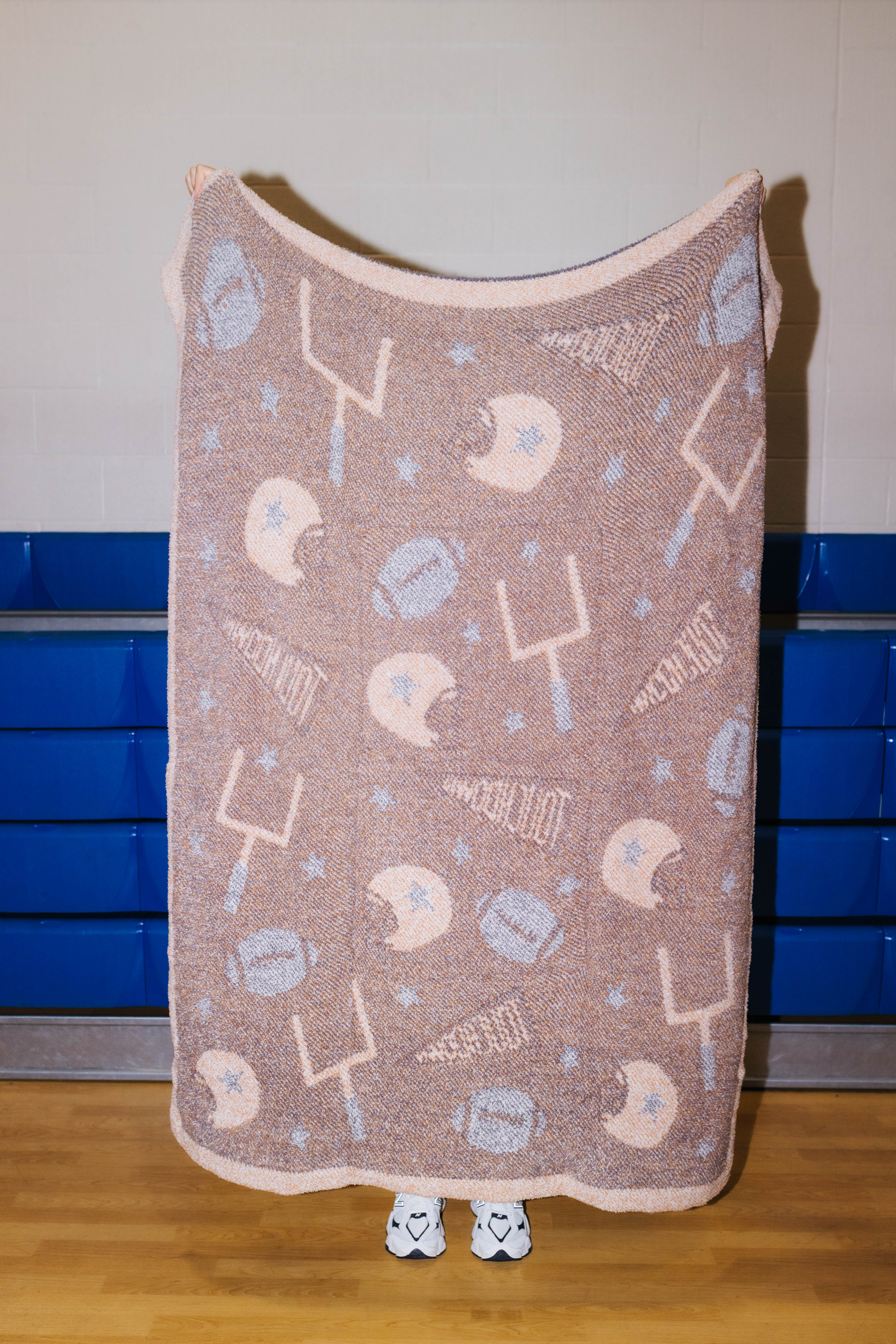 A fleece throw blanket with a football theme, featuring a brown base with lighter graphics of football helmets, footballs, and a football pitch, held up by a person whose feet are visible at the bottom.