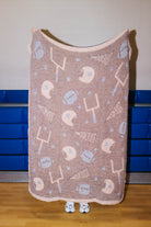 A fleece throw blanket with a football theme, featuring a brown base with lighter graphics of football helmets, footballs, and a football pitch, held up by a person whose feet are visible at the bottom.