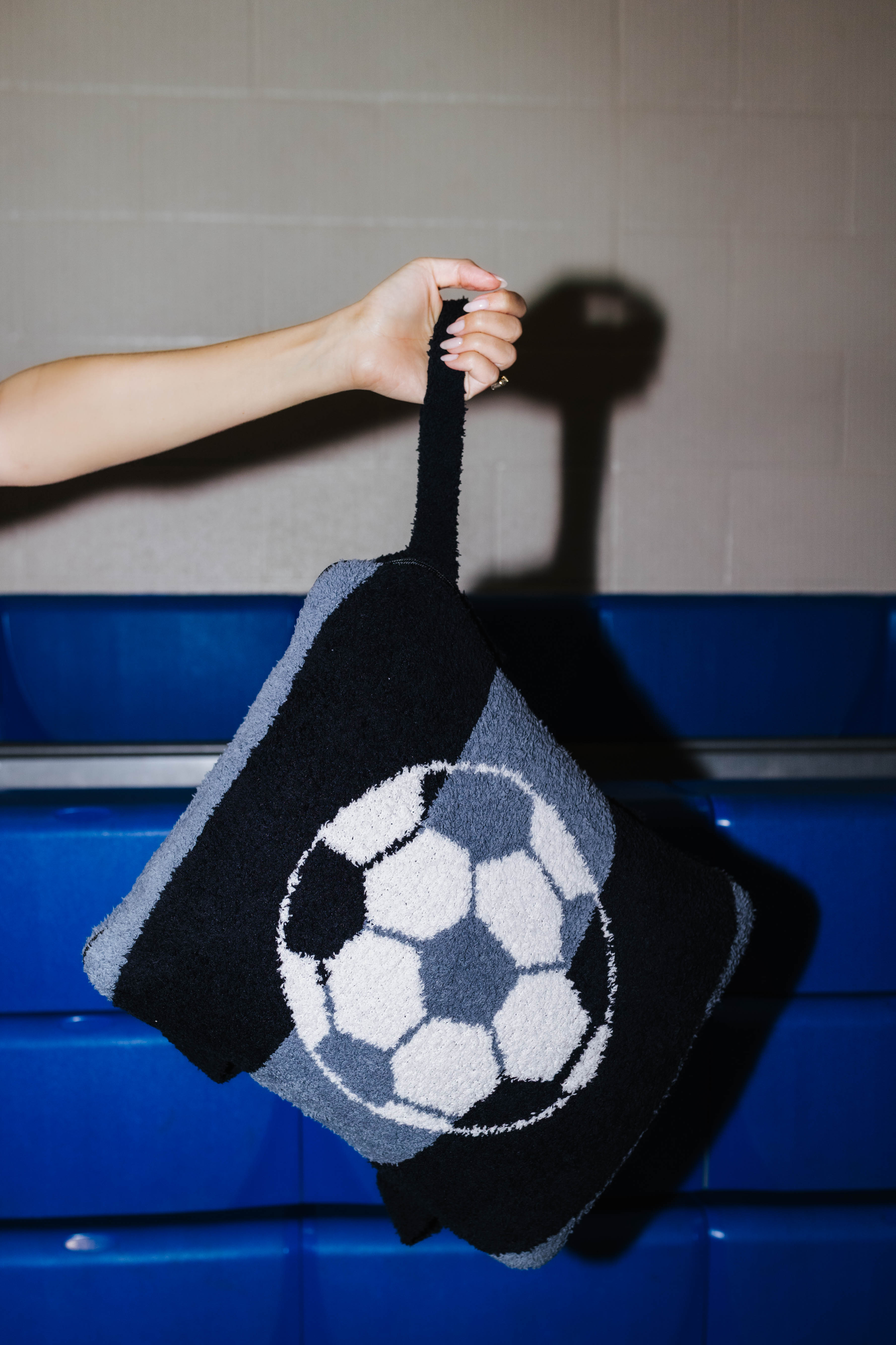 A hand holds a black fuzzy quillow with a large soccer ball pattern on the front, set against a gray tiled wall and blue bench background.