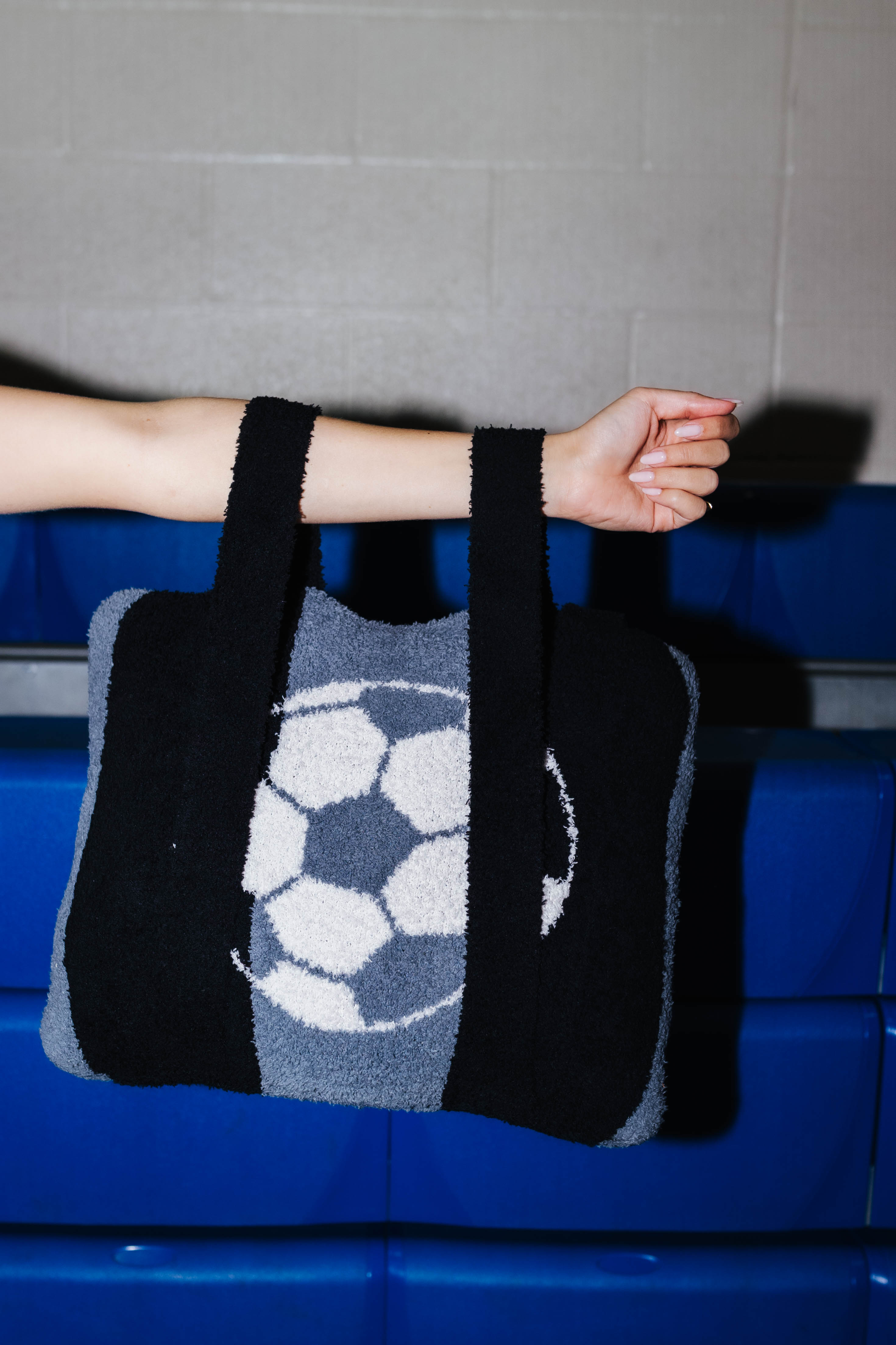 A fuzzy quillow with soccer ball pattern, held by a person's arm against a gray wall and blue padded seating.