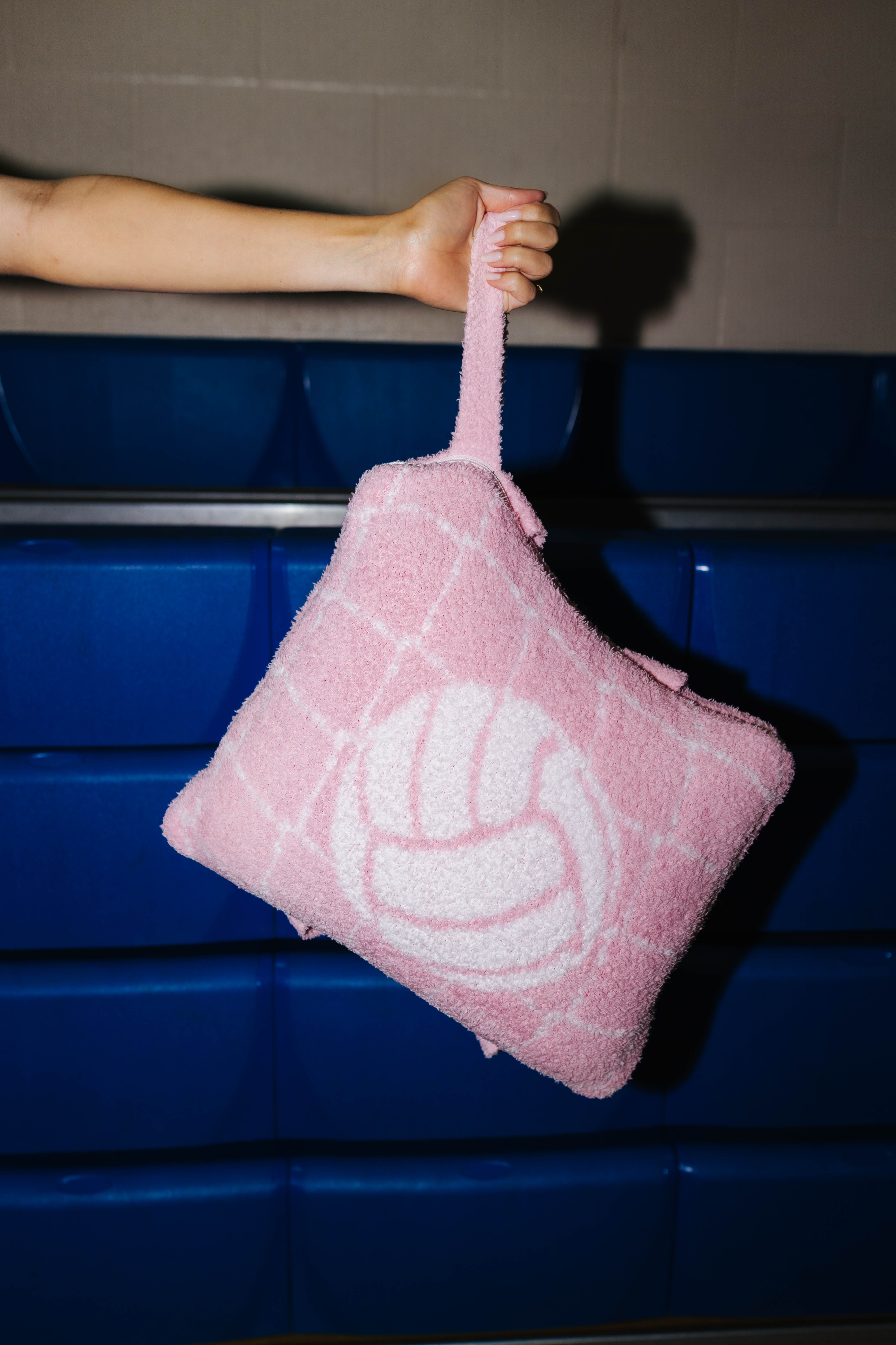 A pink quillow with a volleyball graphic and a top handle, held by a person's arm in front of blue bleachers